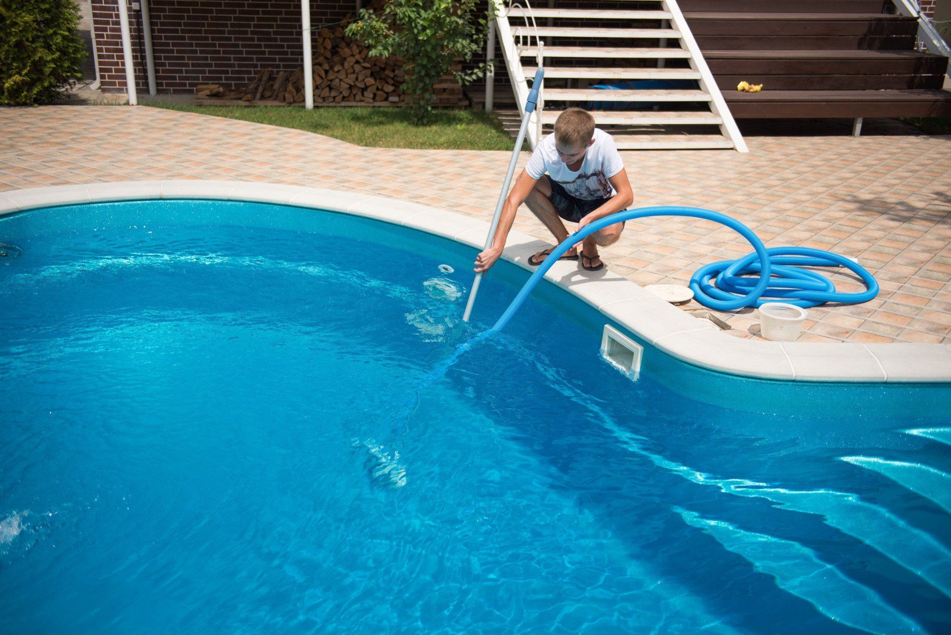 Man cleaning a blue swimming pool with a hose, steps and patio in background.