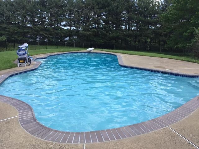 Swimming pool with blue water and surrounding concrete deck. A wheelchair lift is on the side, near the grass.