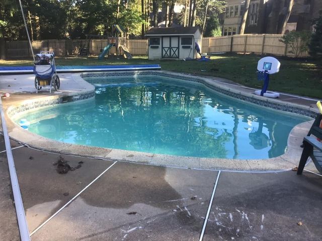 A pool with blue water surrounded by a concrete patio, a fence, and a small wooden shed.