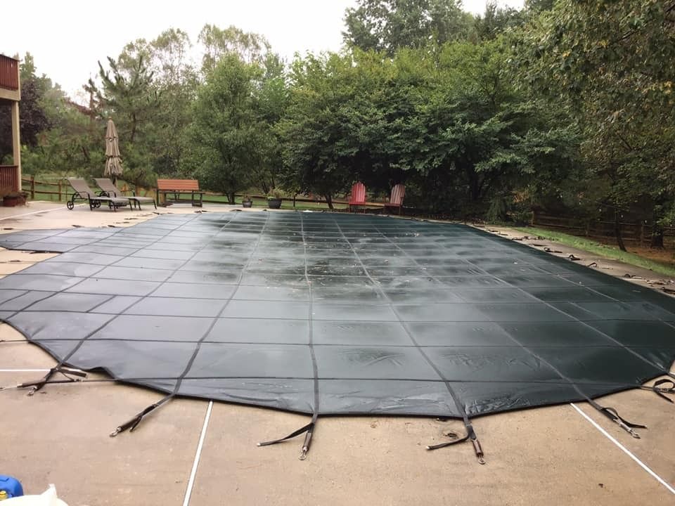 Pool covered with a dark green safety cover, surrounded by concrete patio and trees.