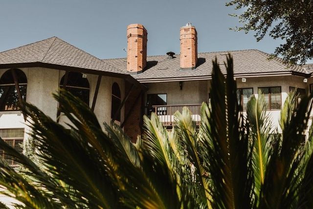 House with tiled roof and two chimneys, partially hidden behind tall palm fronds.
