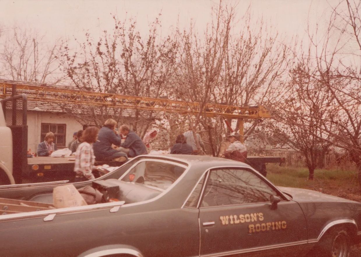 Vintage green car at an outdoor gathering, with people standing nearby under bare trees and a building in the background.