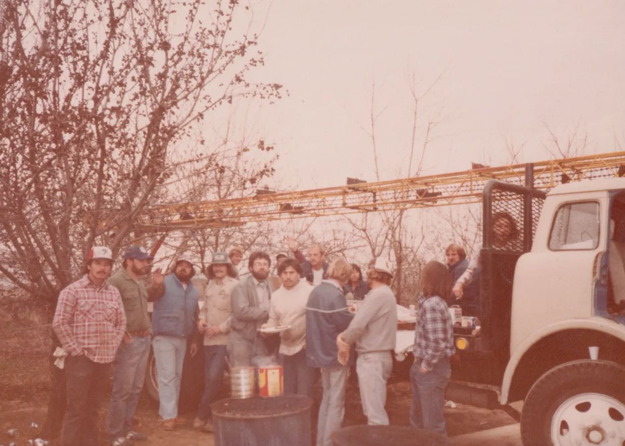 Group of people standing outdoors beside a white truck under bare trees on a cloudy day.