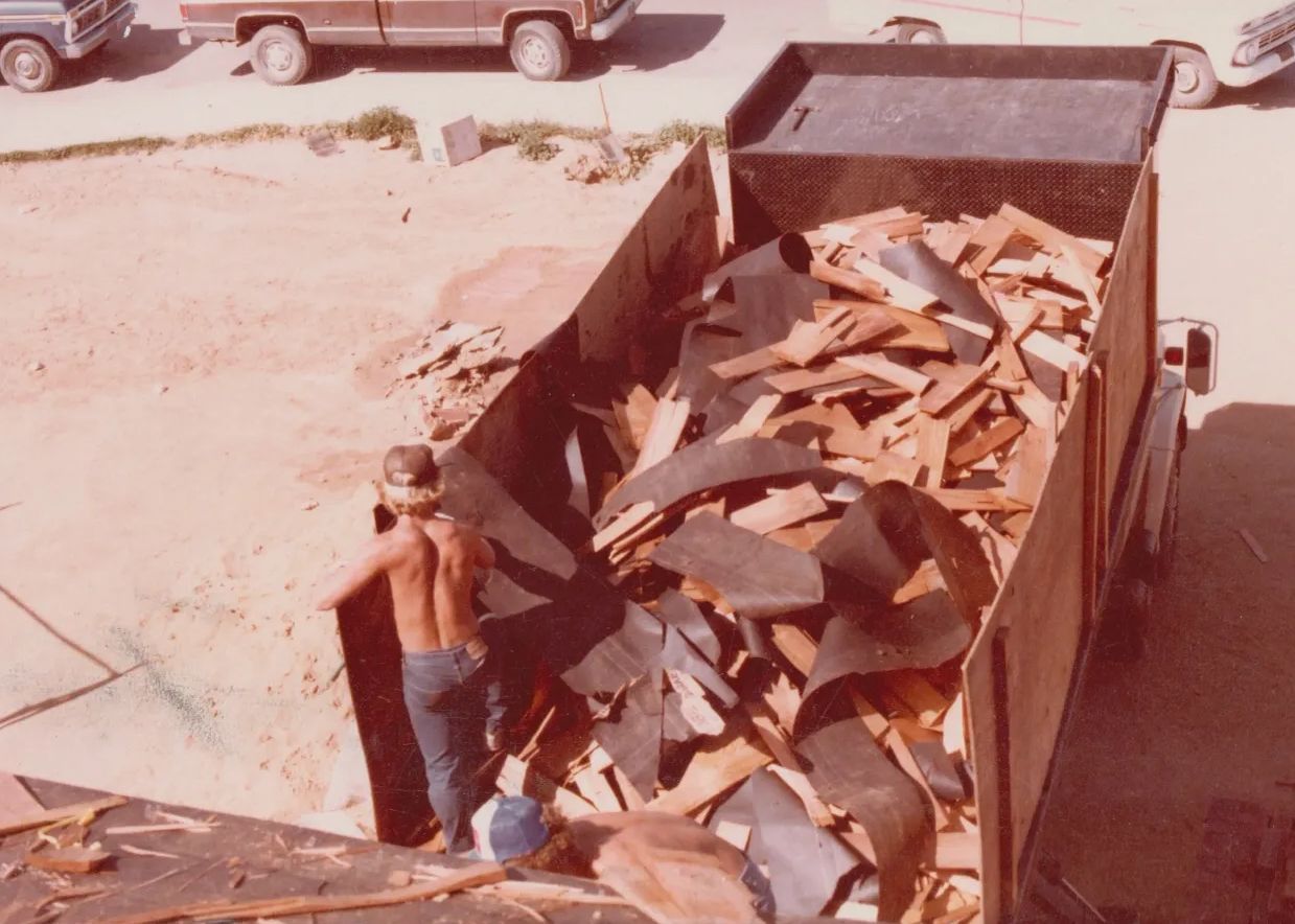 Worker loading scrap wood into a dump truck in a dusty yard.