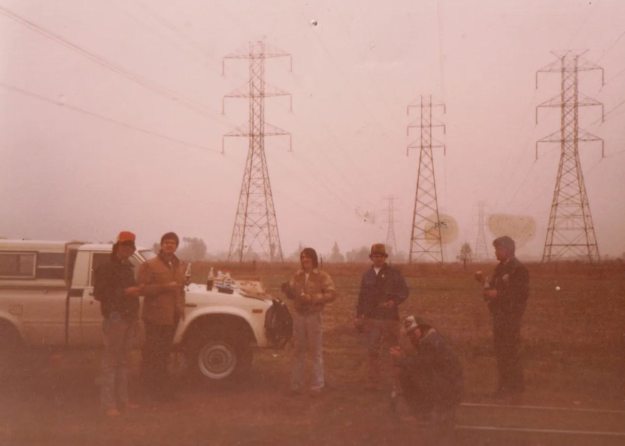 People standing near a white truck in a dusty field with power lines and transmission towers at dusk.