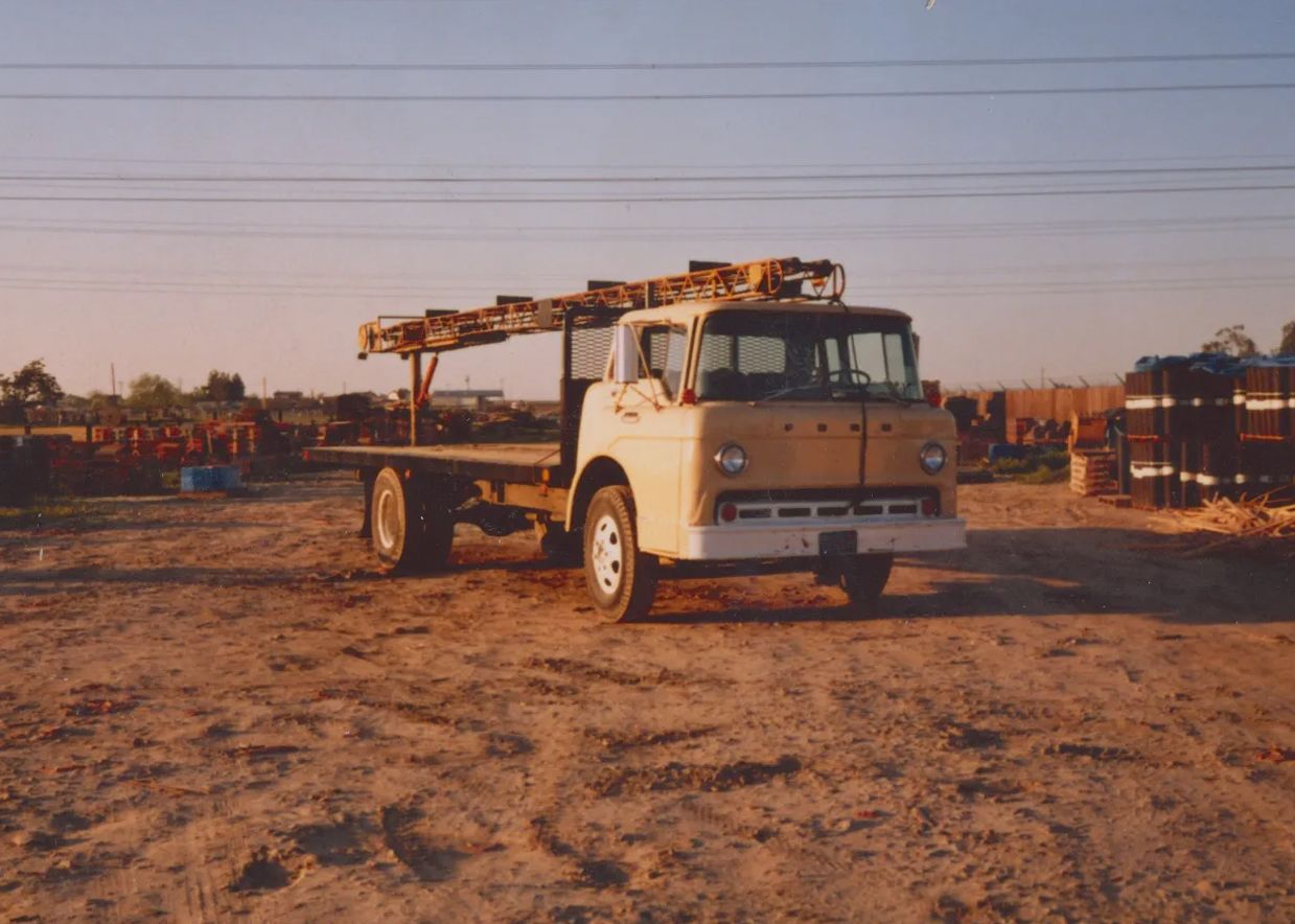 White flatbed truck parked on a dirt lot at sunset with long metal equipment on the bed.