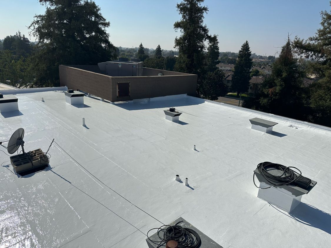 White flat rooftop with vents and equipment, bordered by tall trees under a clear blue sky
