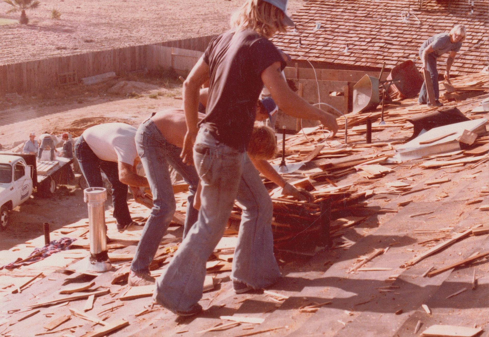 Workers on a roof, prying up shingles with tools amid scattered debris and a ladder nearby.