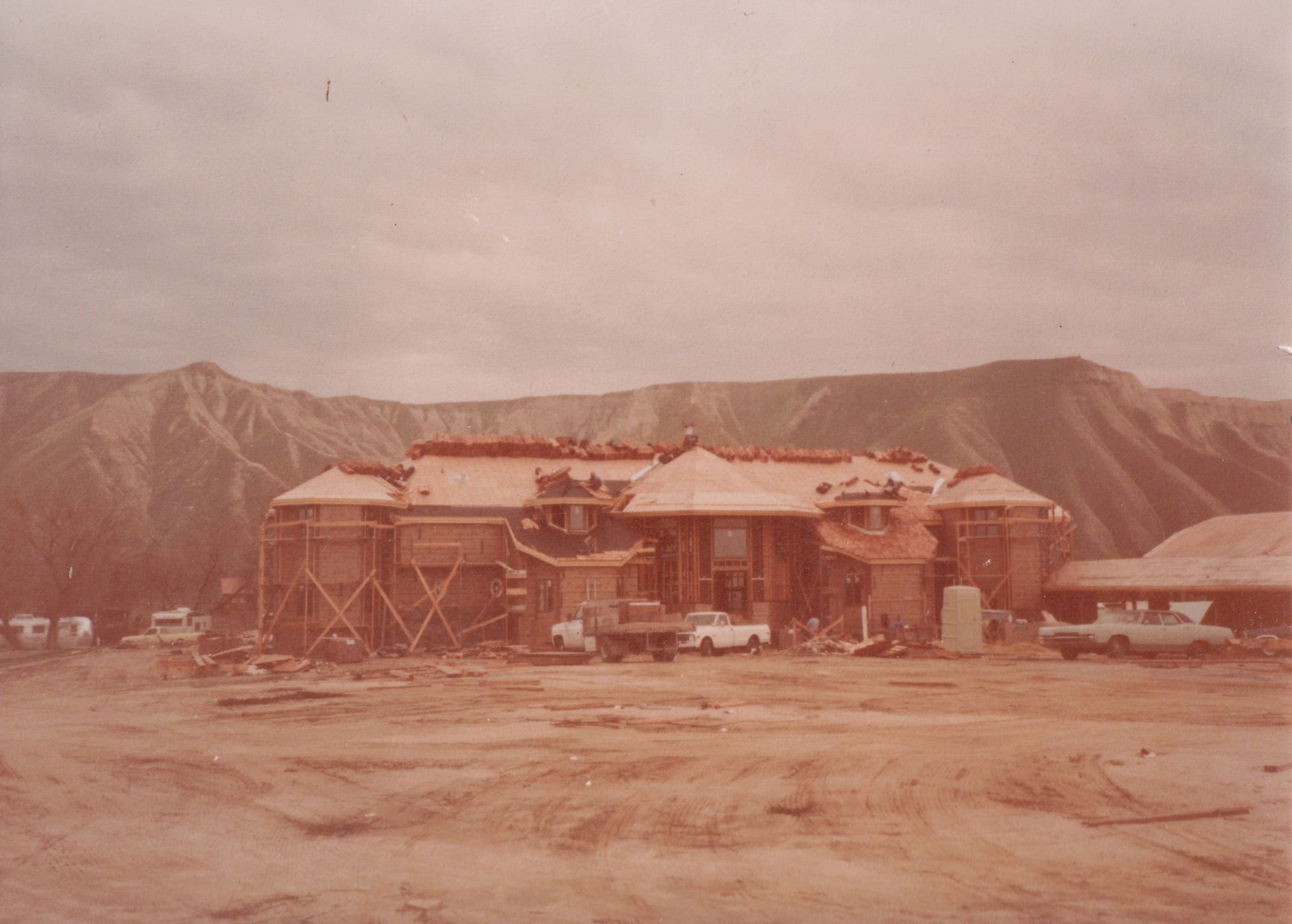 Old desert building under construction with mountains in the background and vehicles parked nearby