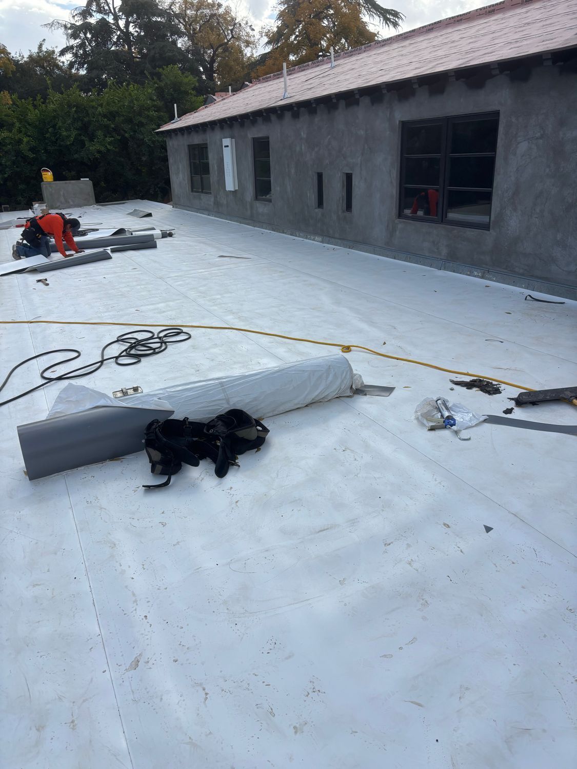Flat white roof under construction beside a gray building, with tools, cables, and workers in the distance.