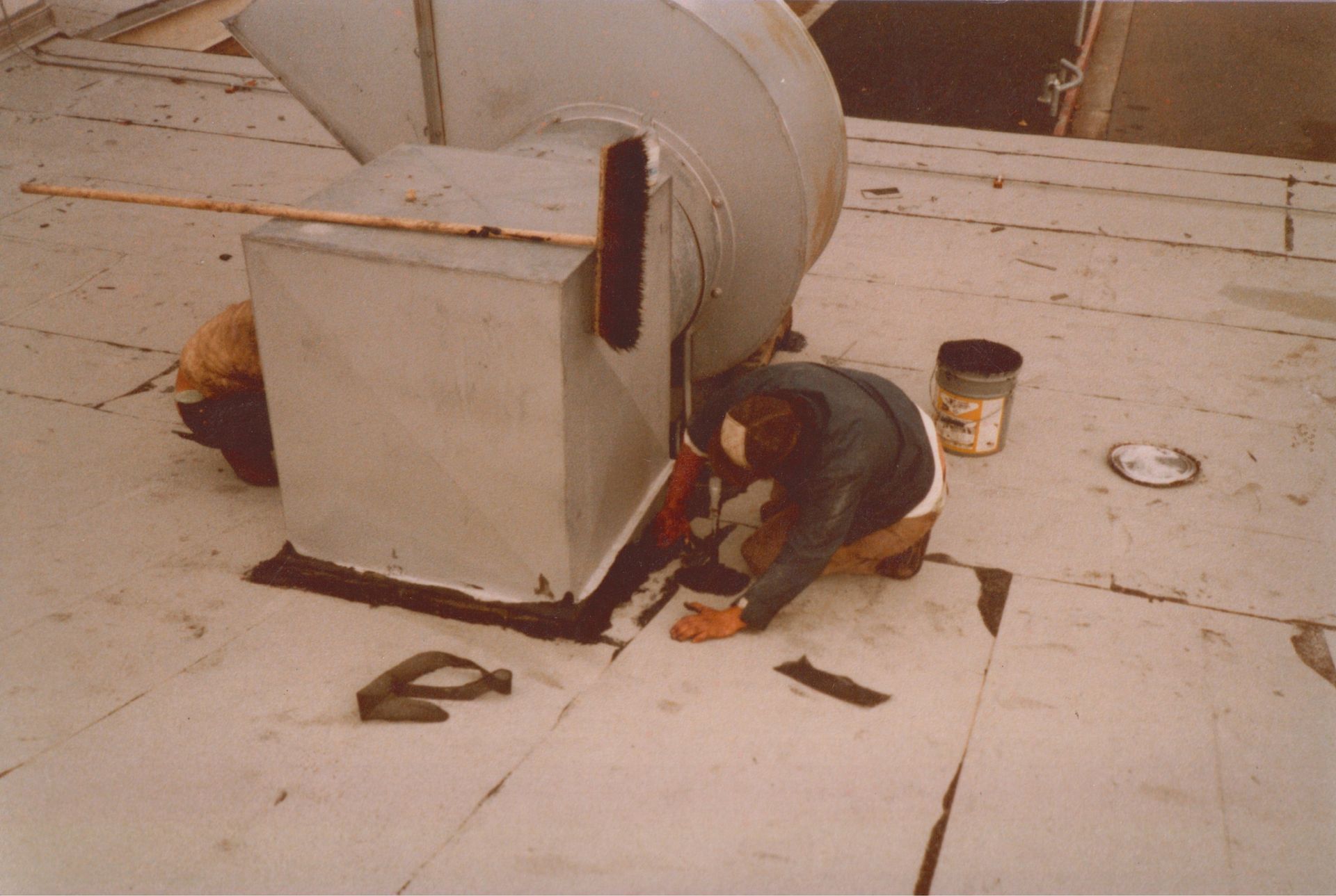Person crouched by a large metal machine on a rooftop, working amid scattered tools and debris.