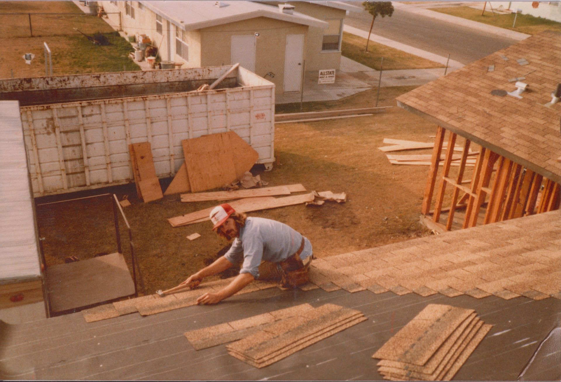Worker laying shingles on a roof at a construction site, with nearby buildings and lumber stacks.