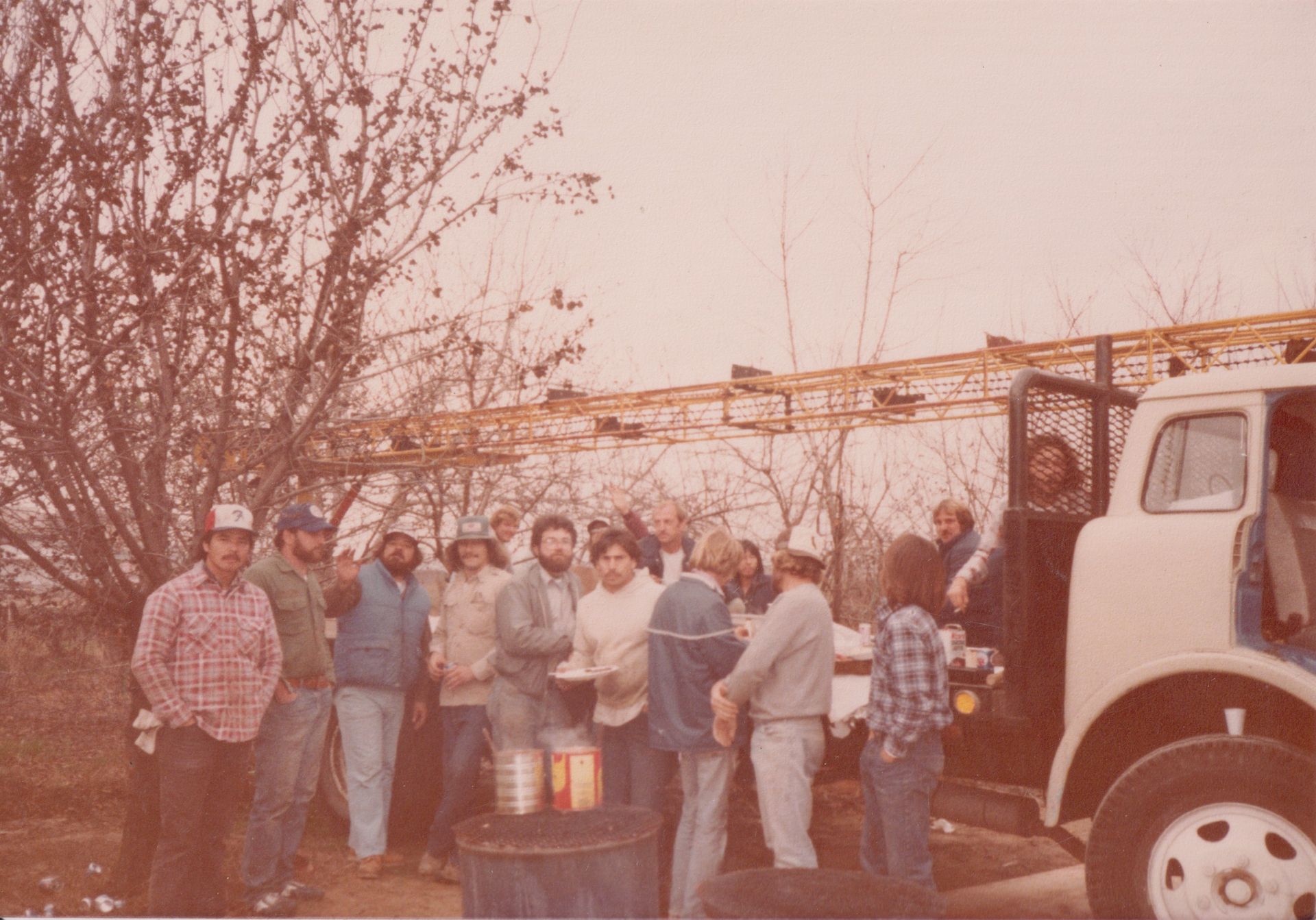 People standing outdoors beside a truck near bare trees and a barrel in a dusty yard