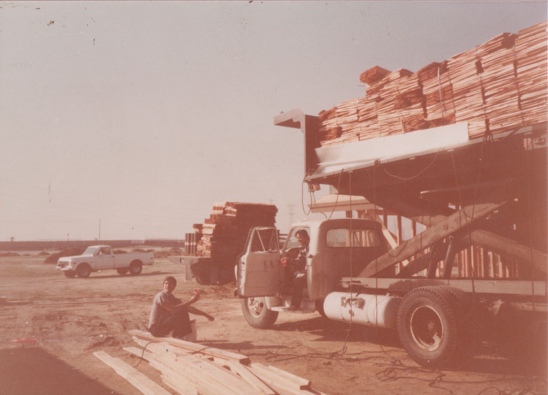 Forklift loading stacks of lumber onto a truck in a dusty yard, with a worker nearby.