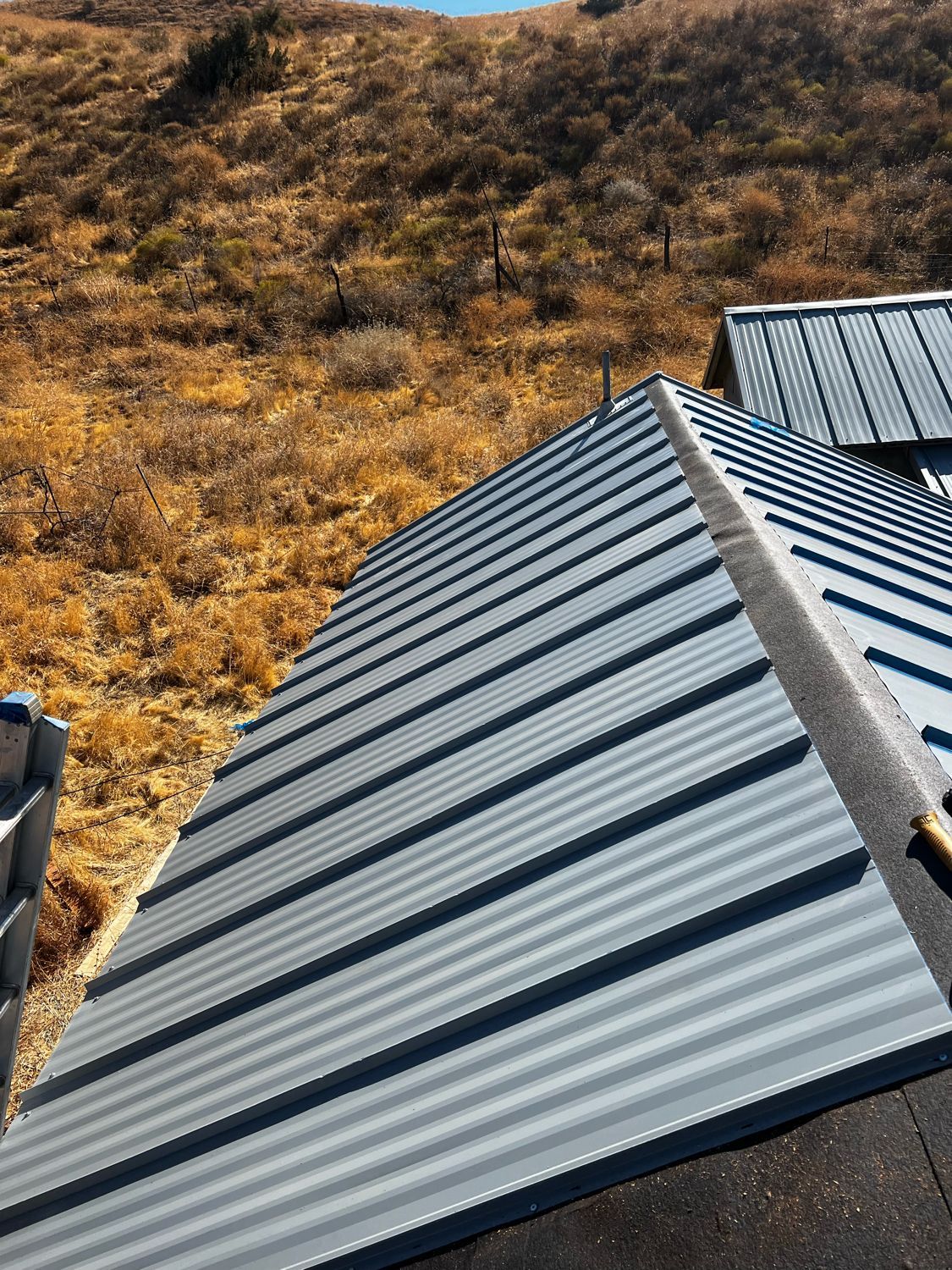 Metal roof panels on a hillside building with dry grass and trees in the background