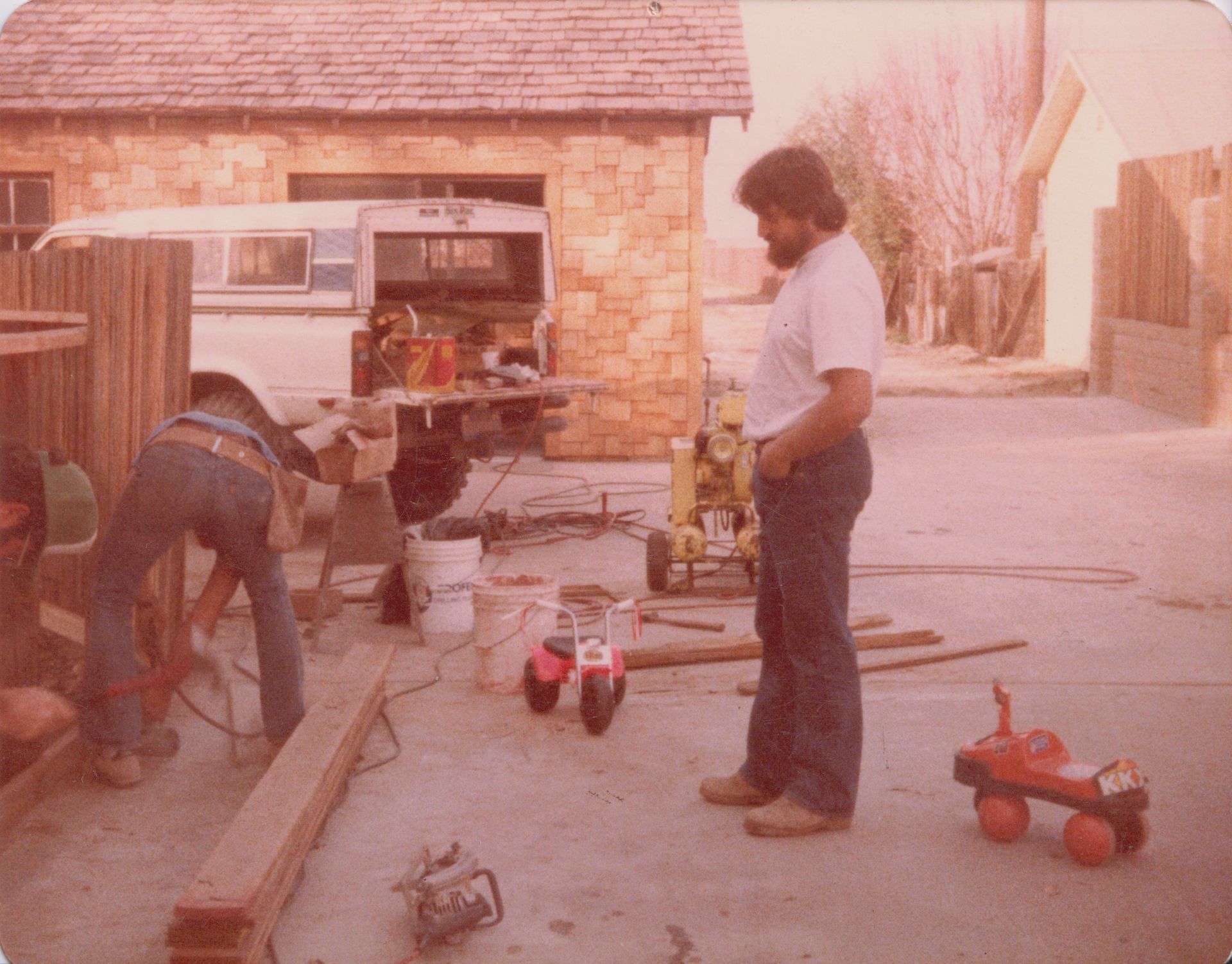 Two people working on a driveway, with tools and toy cars near a brick garage.