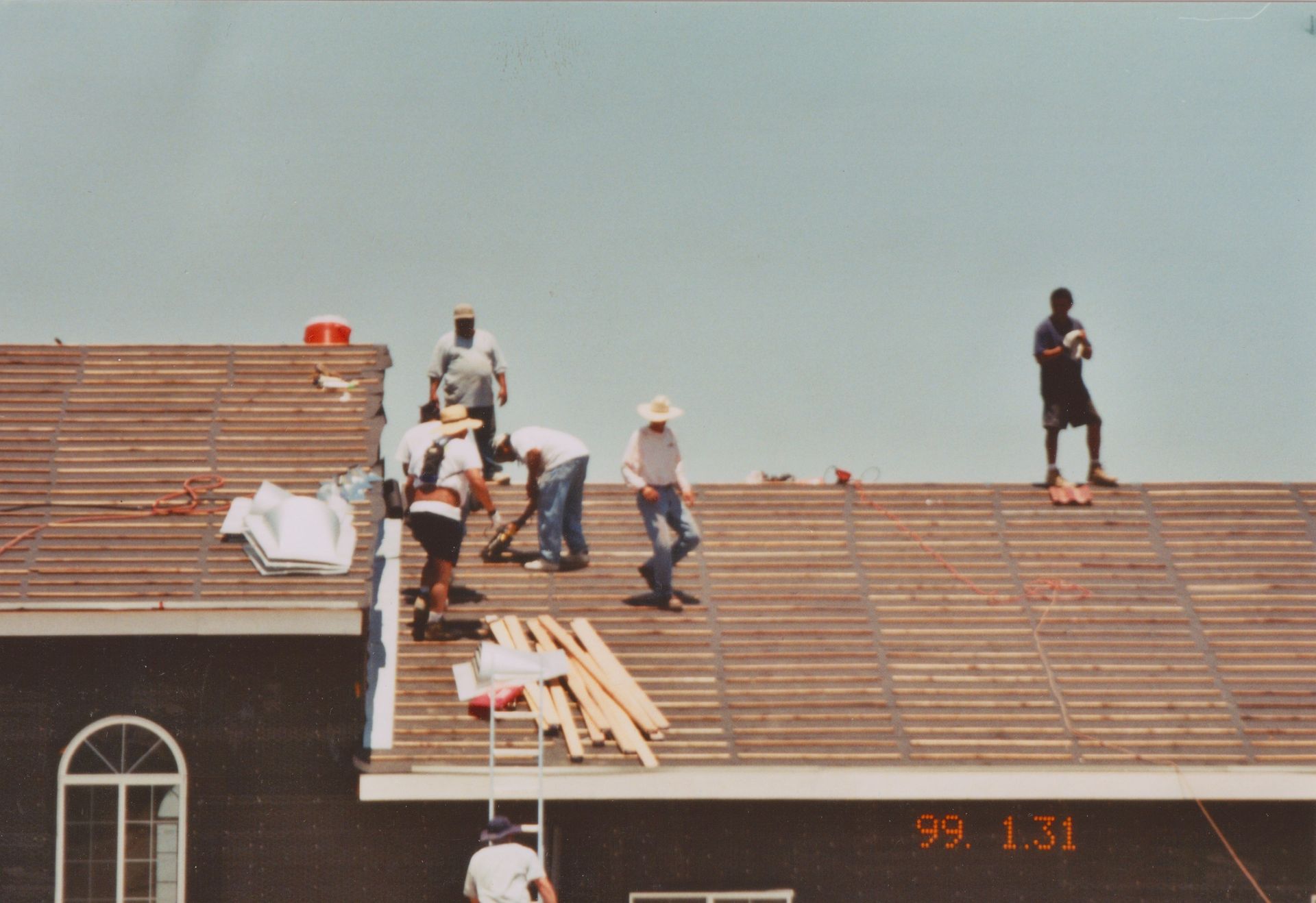 Workers on a sloped roof installing shingles, with stacked materials and a clear sky in the background.