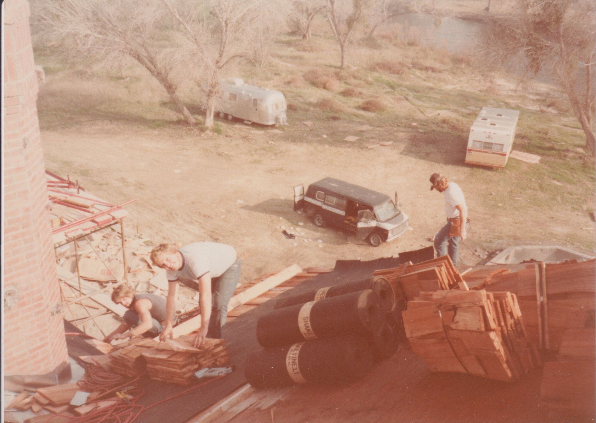 Men repairing a roof beside a car and tree in a dusty yard