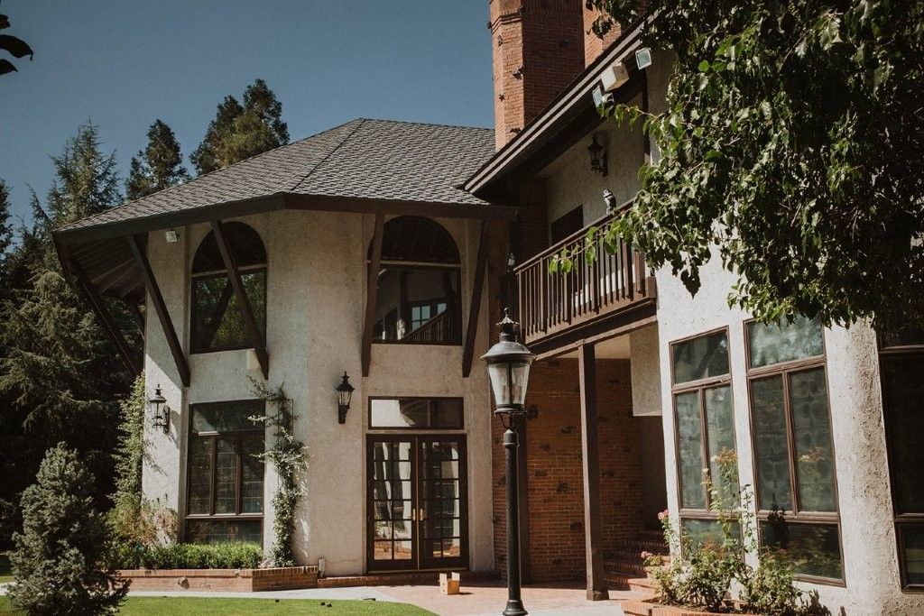 Large stucco house with a tiled roof, brick chimney, and white-trimmed windows amid trees and lawn