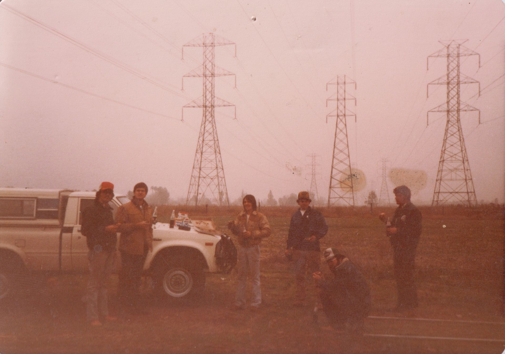 People standing beside a white truck near power lines in a dusty field at dusk