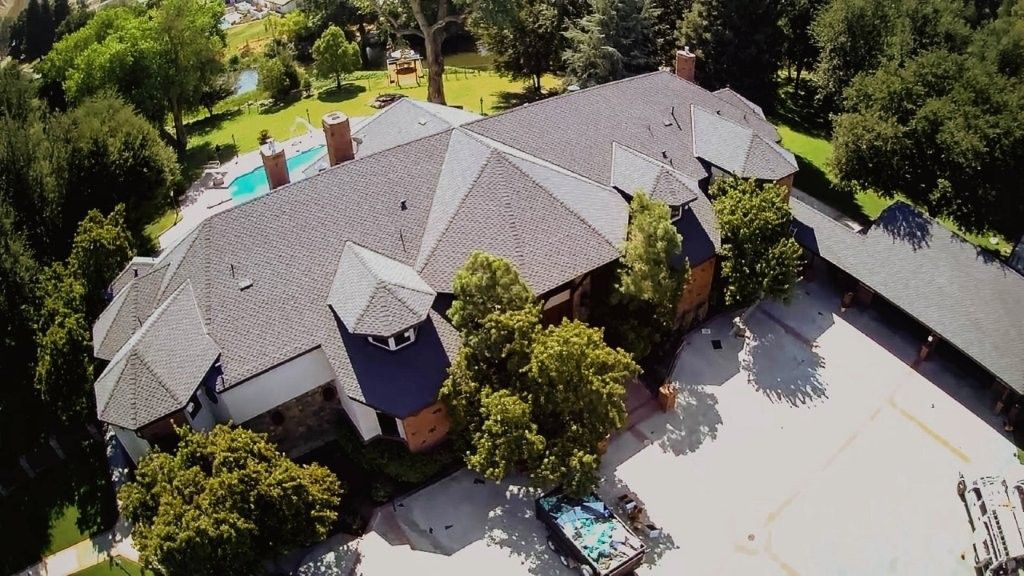 Aerial view of a large gray-roofed house with a pool, driveway, and trees surrounding it