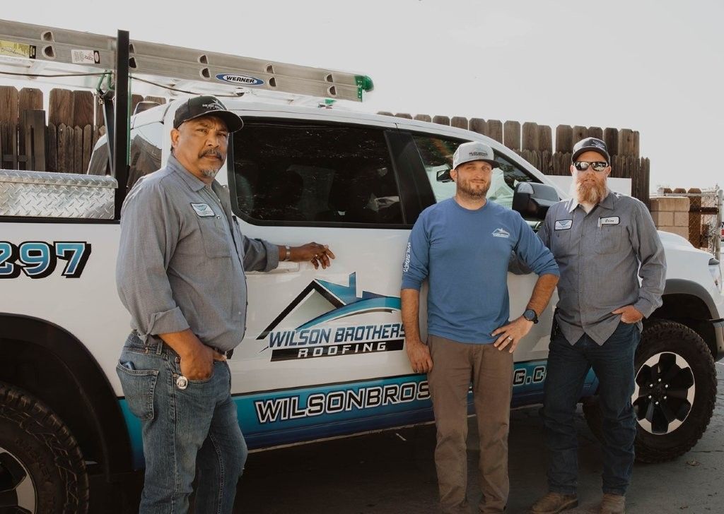 Three workers standing beside a white utility truck with ladder racks and company decals outdoors