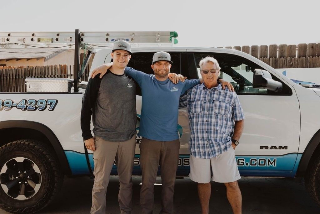 Three people standing arm-in-arm beside a white pickup truck in front of a fence.