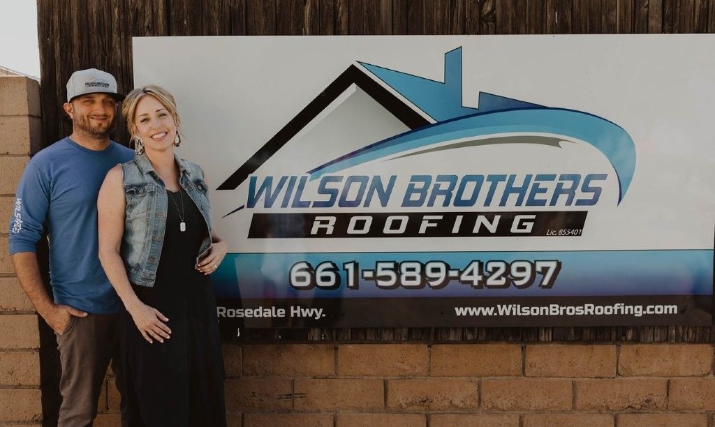 Two people stand beside a Wilson Brothers Roofing sign with logo, phone number, and website on a brick wall.