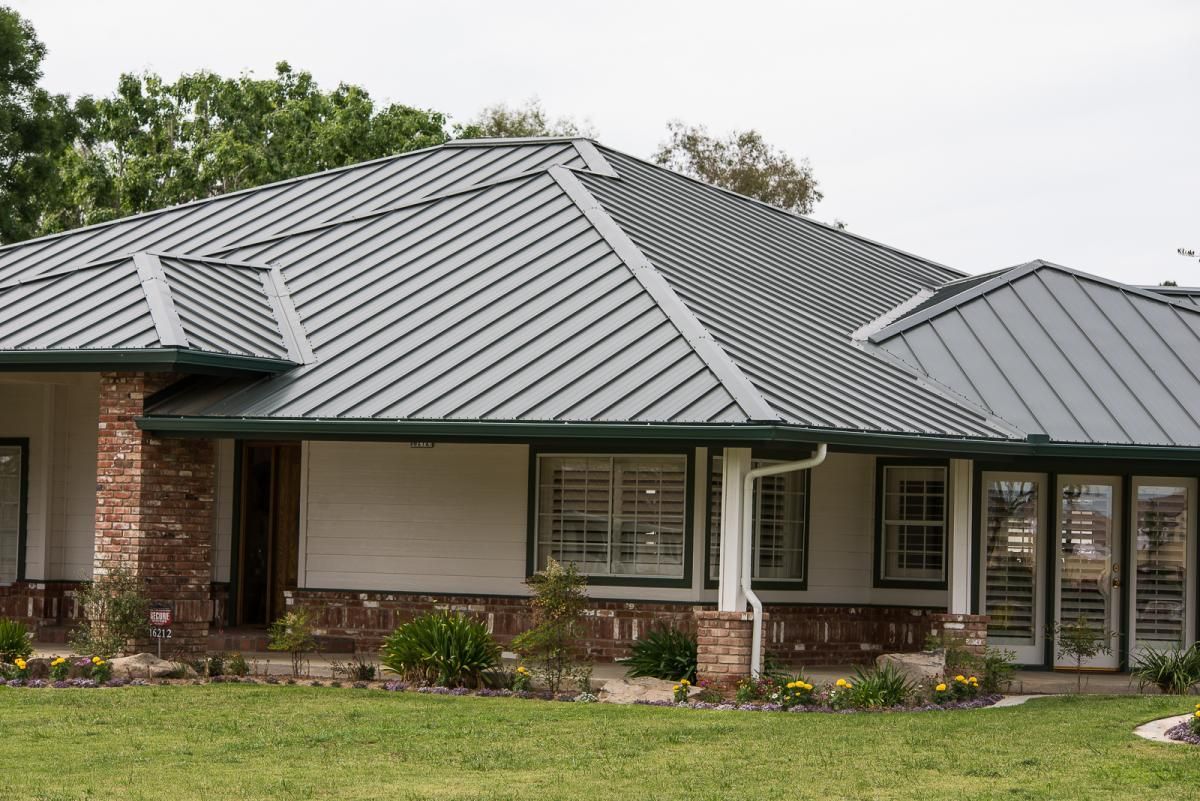 Single-story brick house with a gray metal roof and front porch surrounded by lawn and shrubs