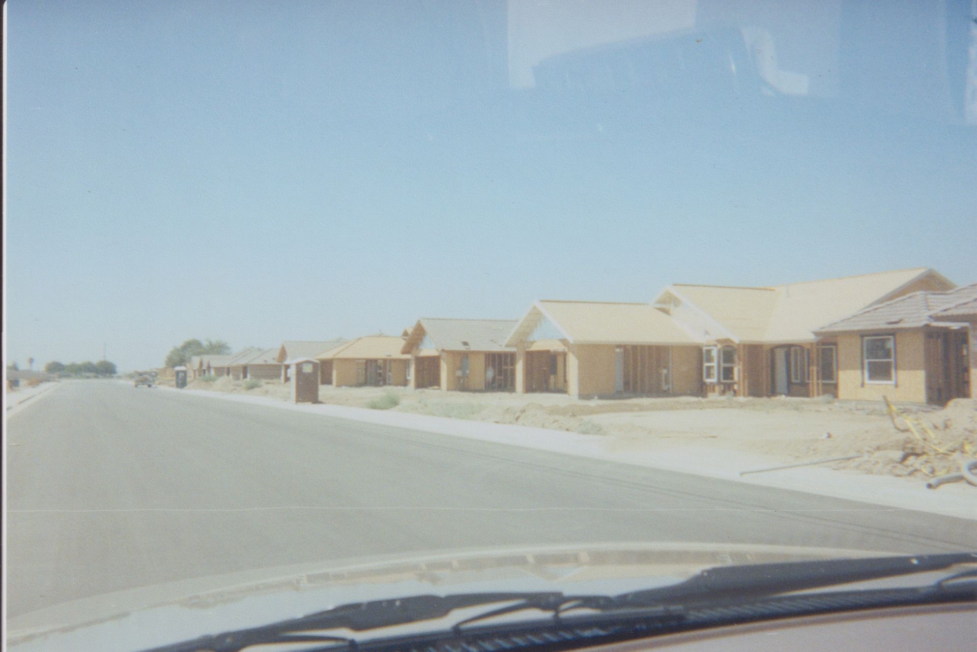 A dusty suburban street with rows of beige houses under a clear blue sky, viewed from a car windshield.