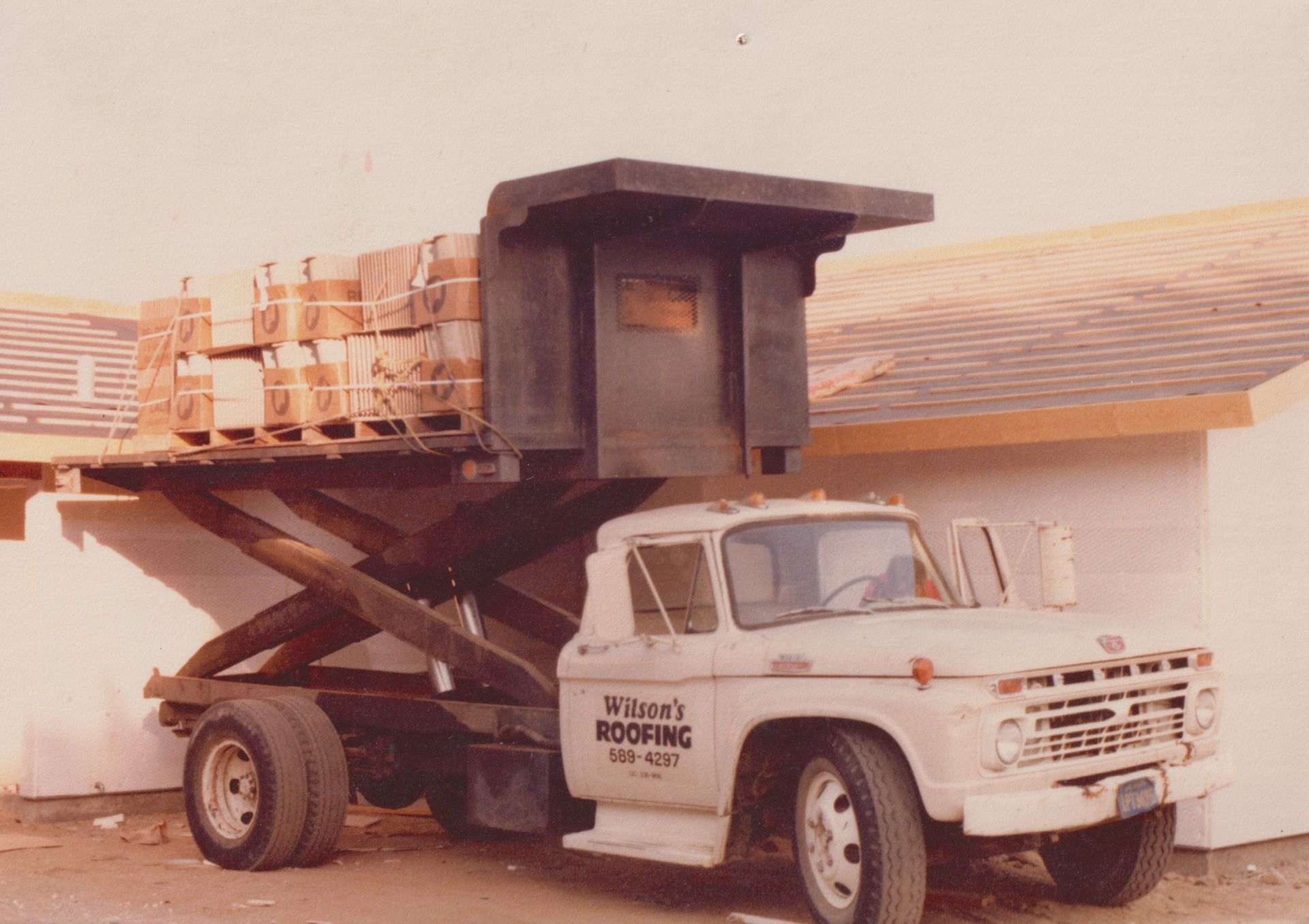 White pickup truck with a raised dump bed unloading stacked crates beside a building.