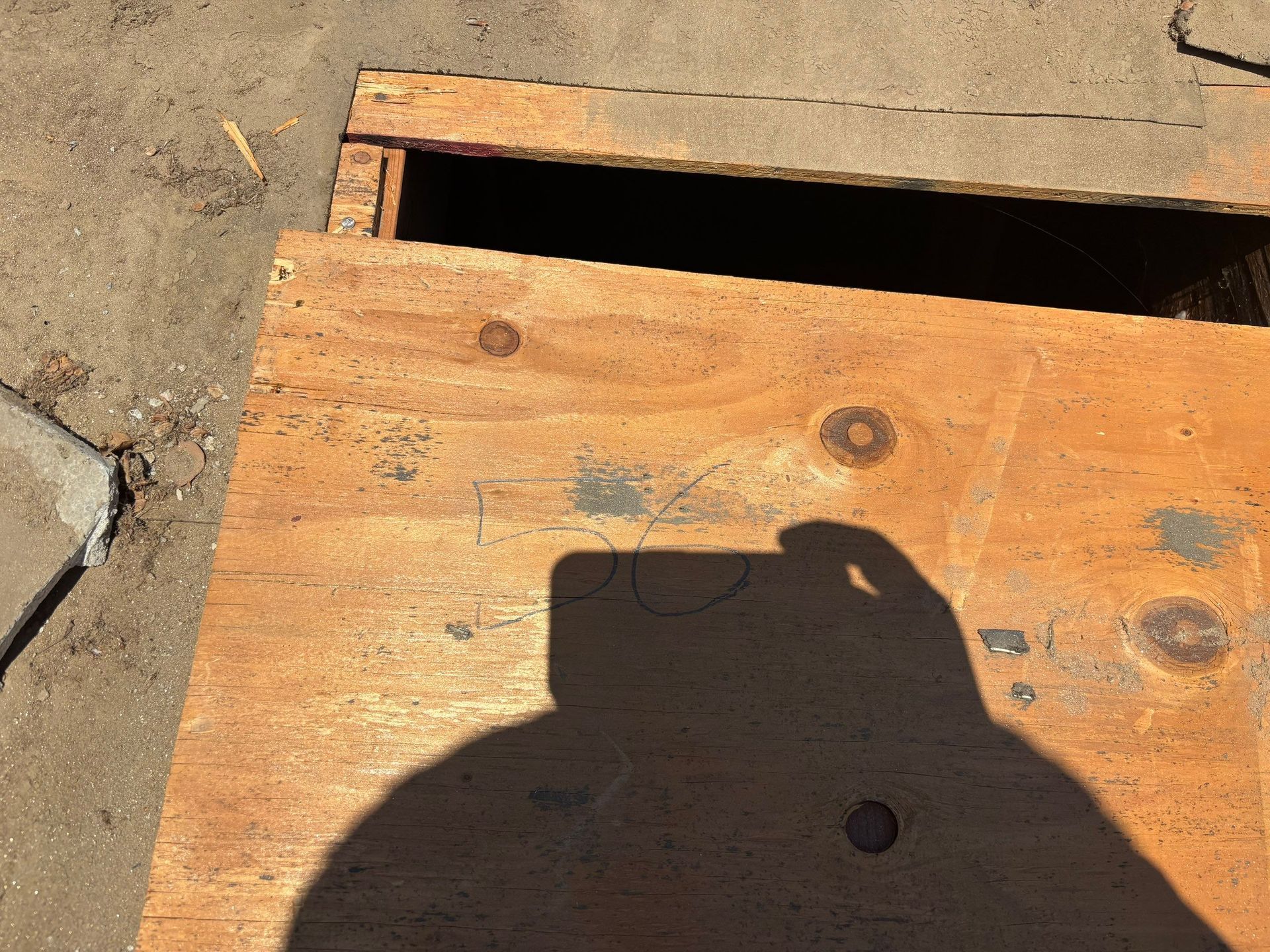 Wooden board covering a dark opening in sandy ground, with a shadow across it.