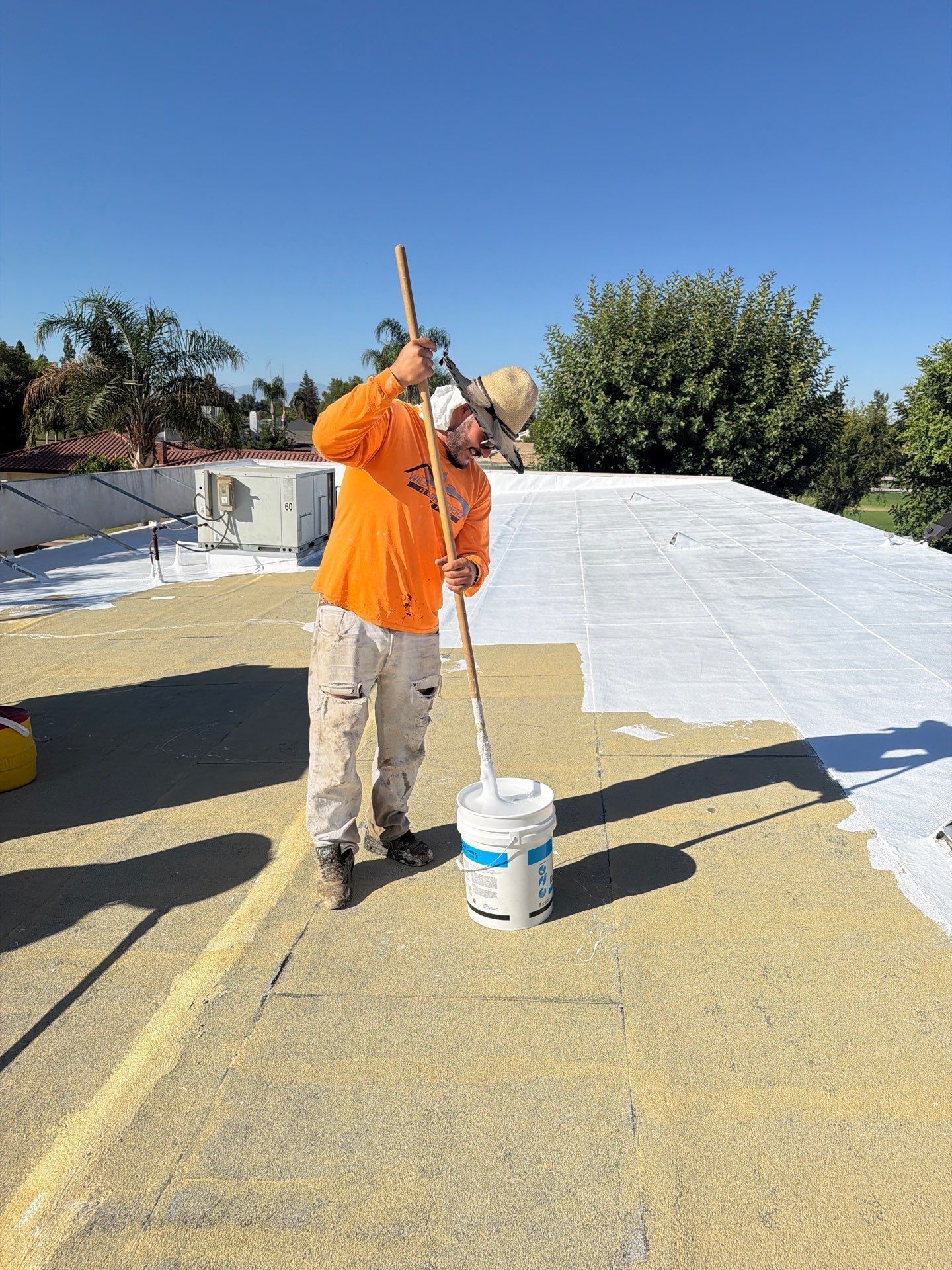 Worker in orange shirt spraying white coating on a rooftop beside a paint bucket.