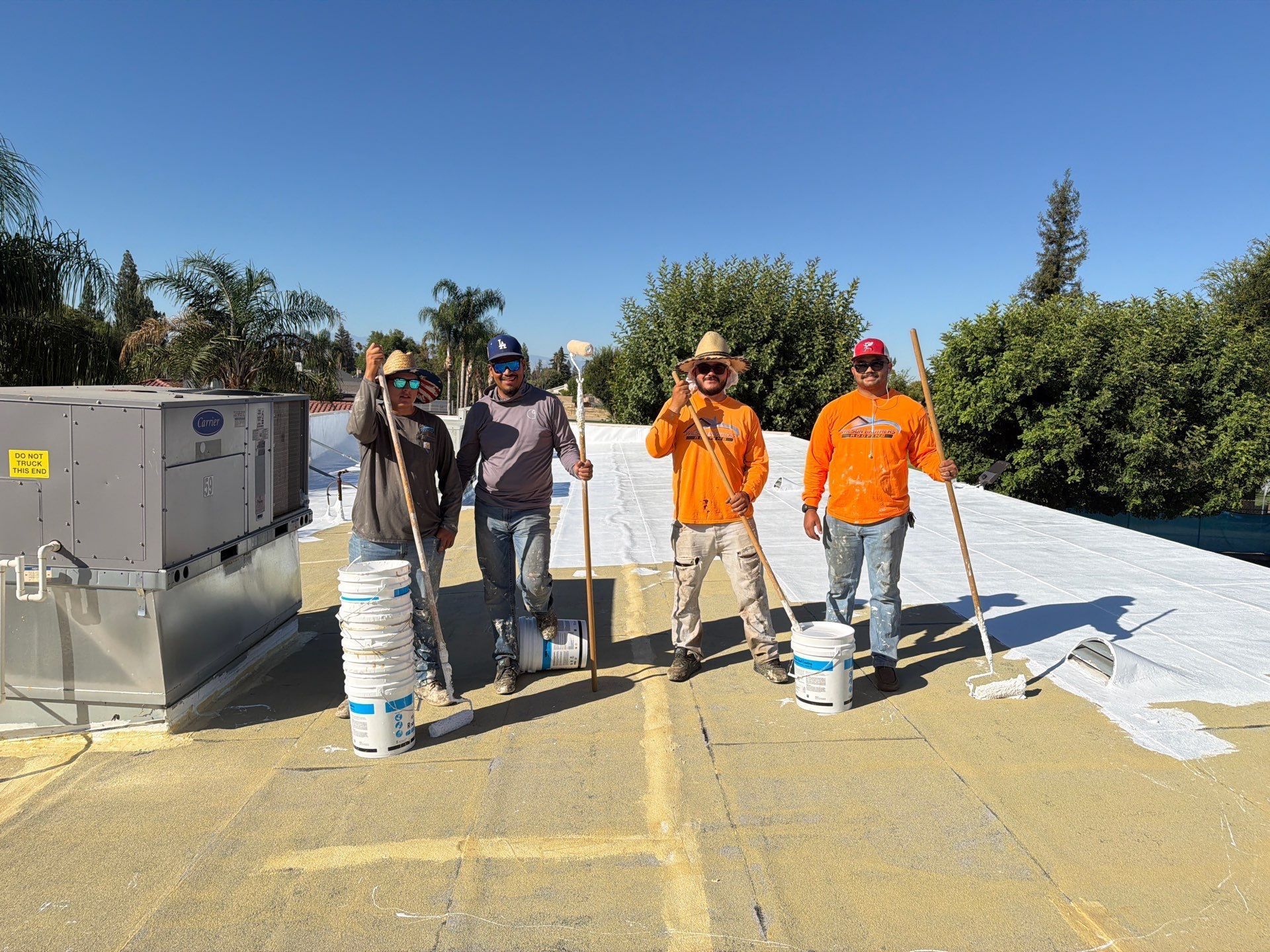 Four workers painting a flat rooftop, with white coating and equipment under a clear blue sky