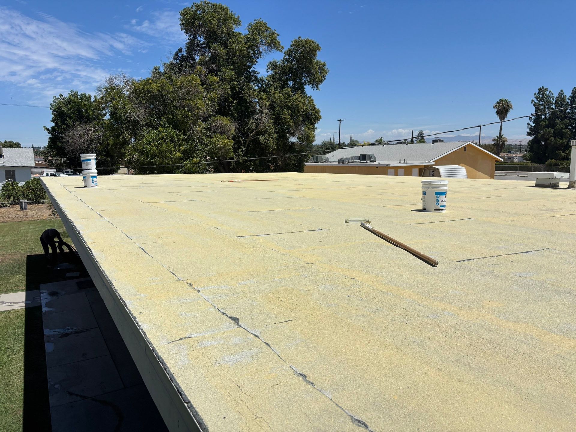 Flat yellow rooftop with vents and a long tool, trees and houses in the background under a blue sky