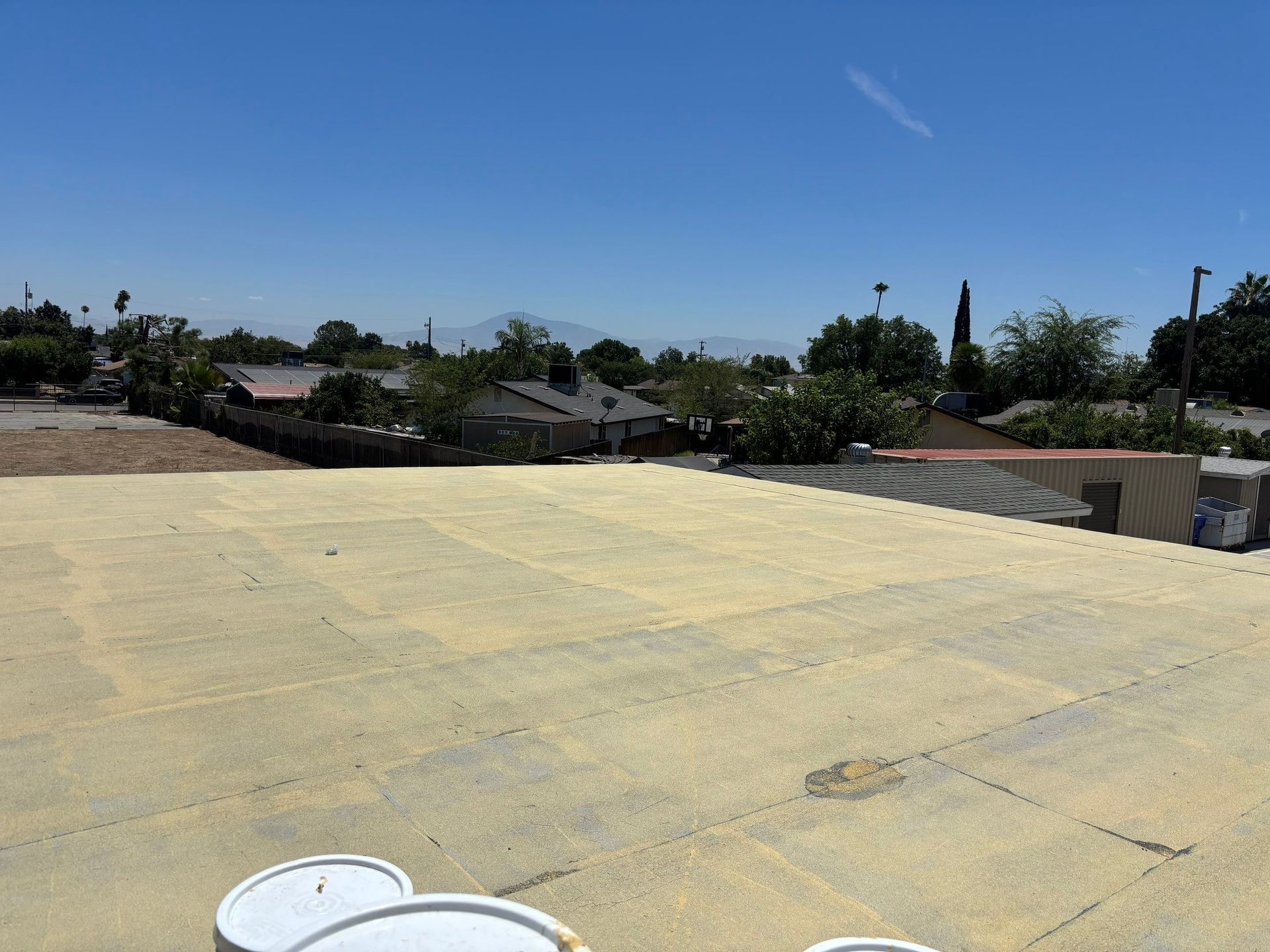 Flat rooftop with white buckets in the foreground under a clear blue sky, overlooking houses and trees.