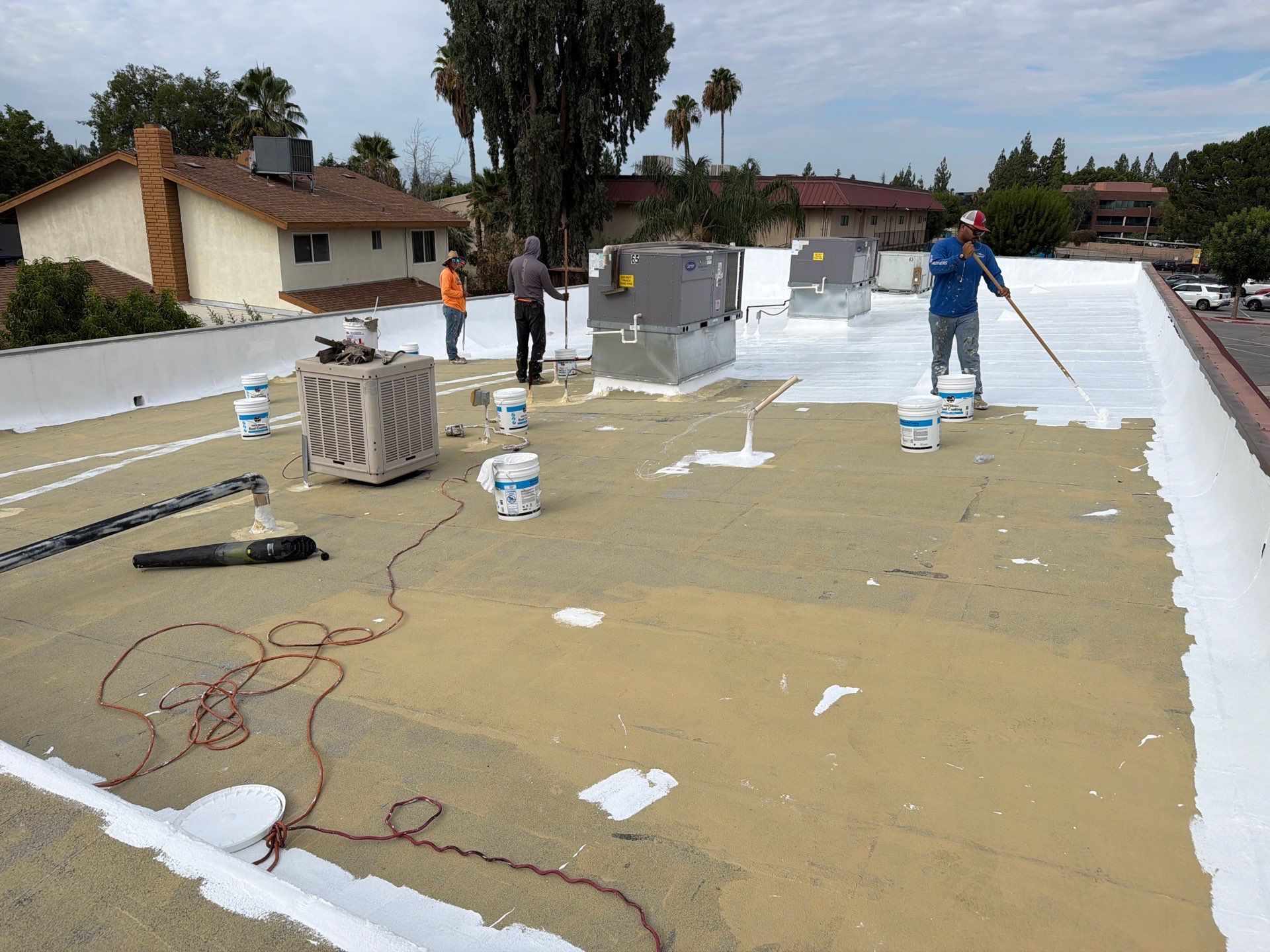 Workers applying white coating on a flat rooftop under construction, with buckets and tools scattered around.