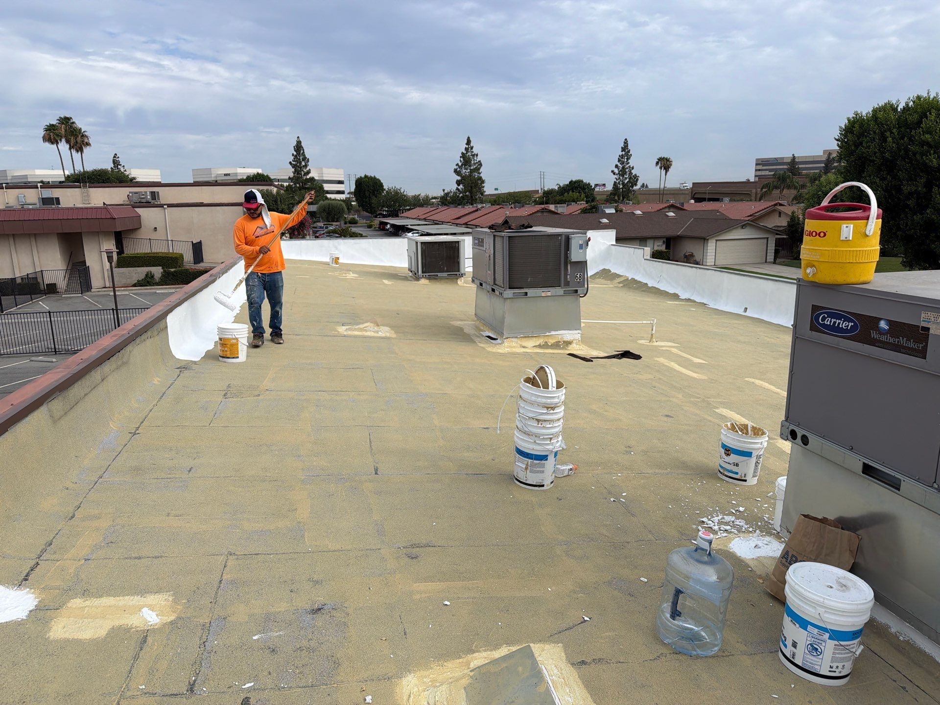 Worker in orange safety gear on a flat rooftop with buckets and tools under a cloudy sky