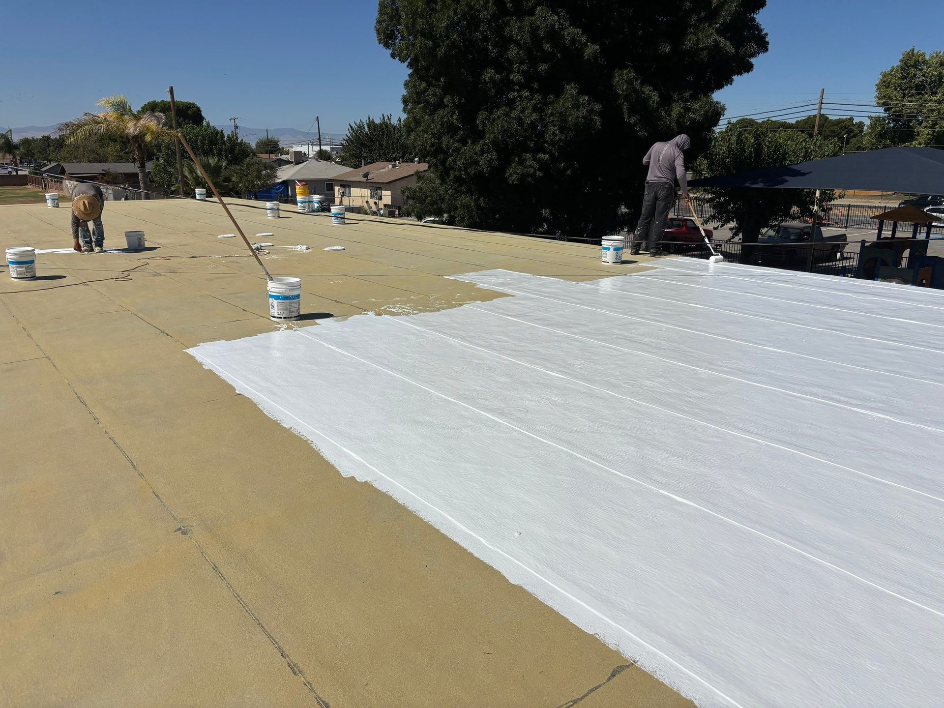 Workers painting a large rooftop white on a sunny day, with buckets and tools spread across the surface.