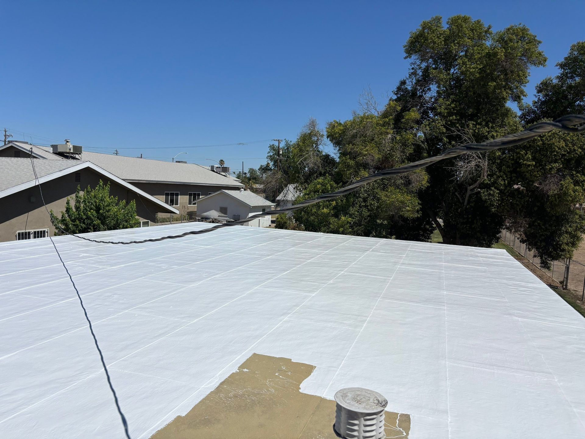 White roof coated in sealant with a bucket in the foreground and trees and houses in the background.