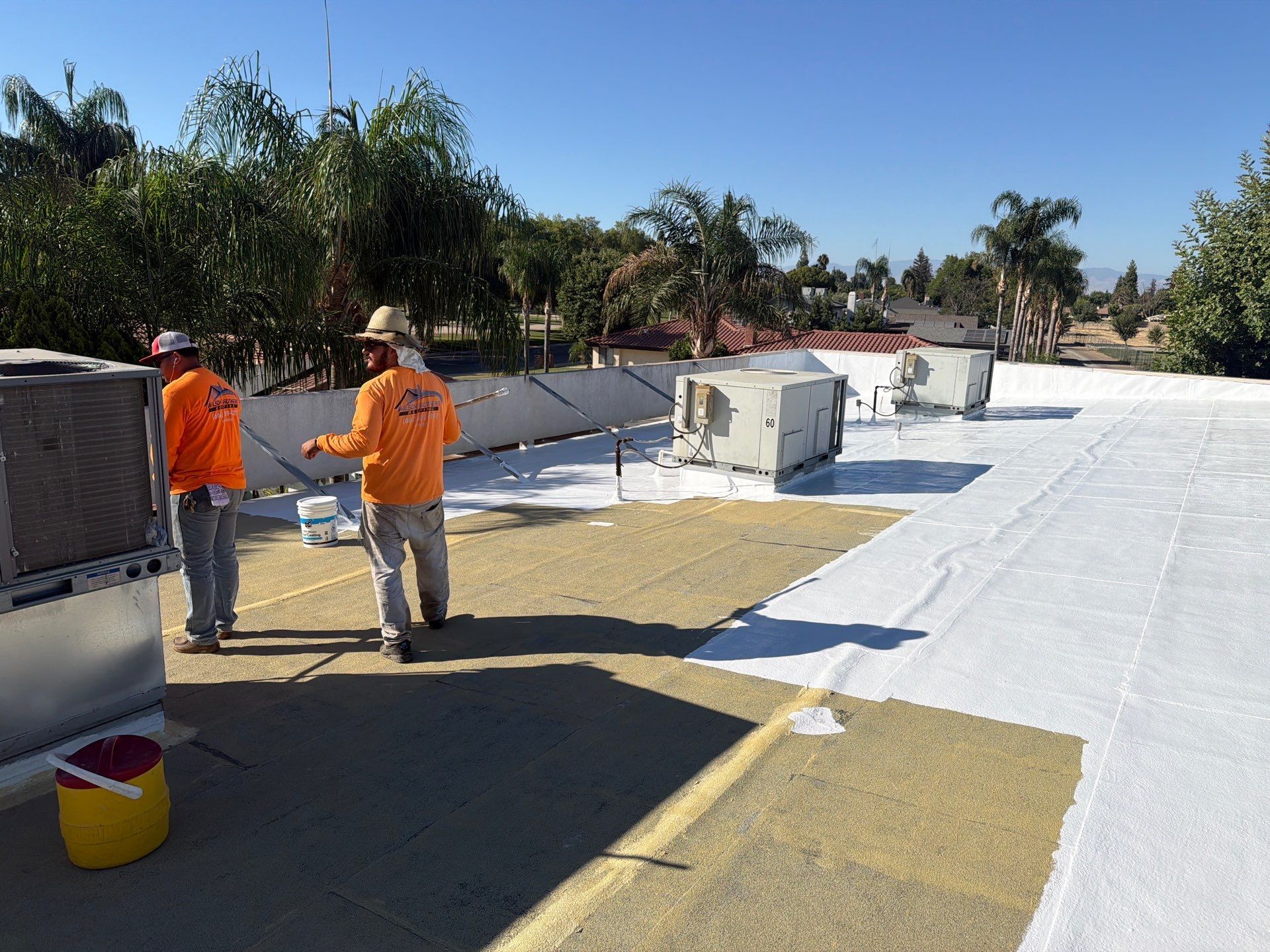 Workers in orange vests applying roofing material on a flat roof in bright sunlight