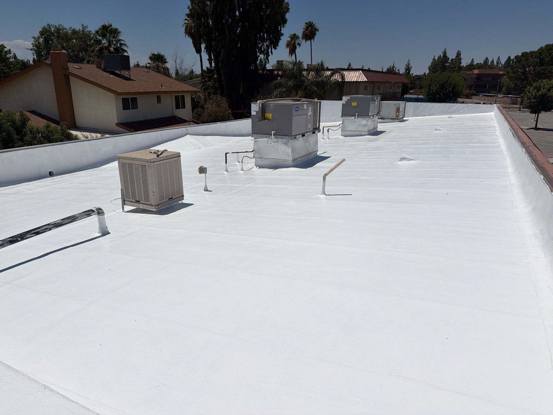 White rooftop with HVAC units and vents under a sunny sky, with houses and palm trees in the background