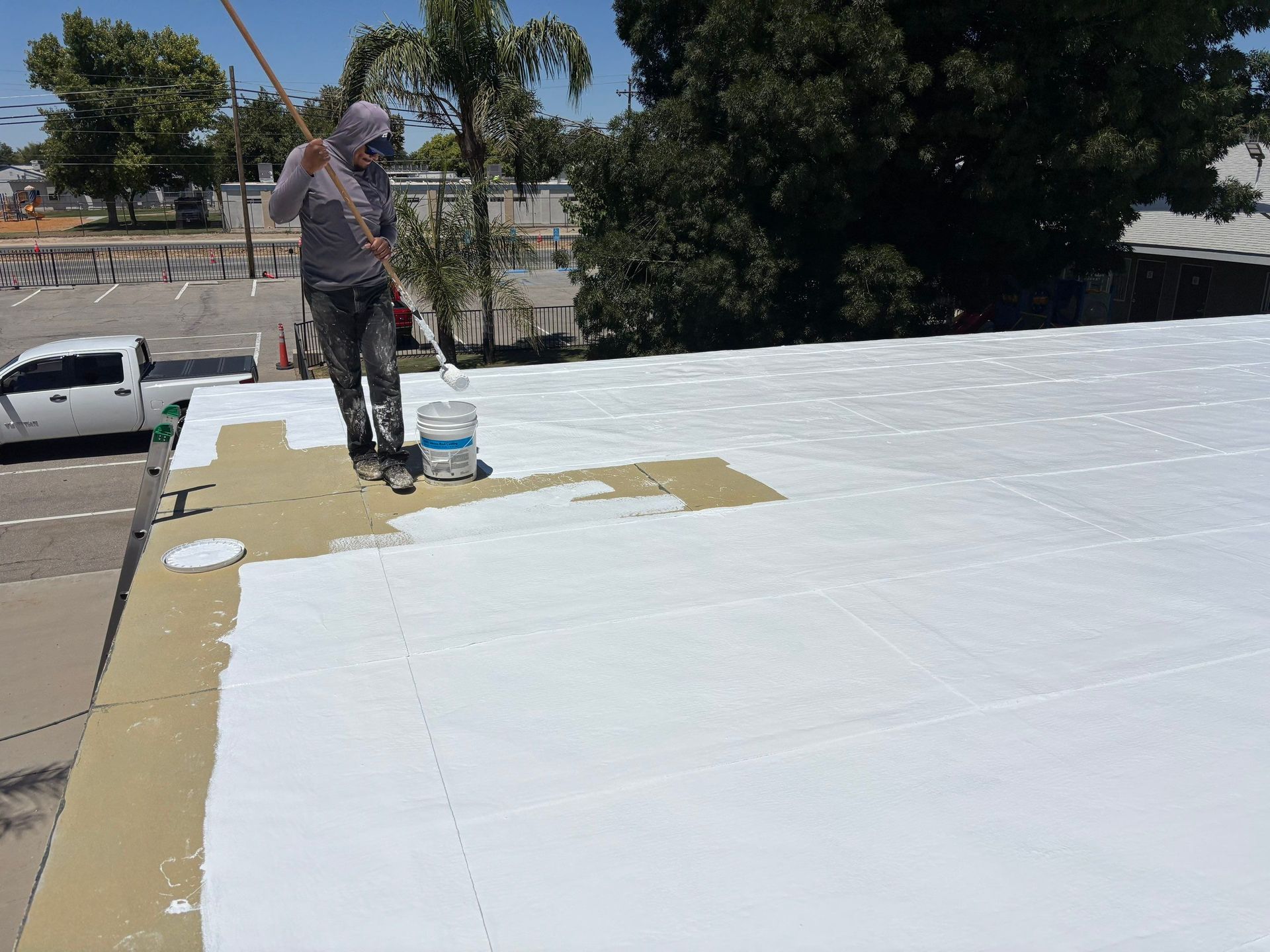 Worker sealing a flat white roof beside trees and parked vehicles on a sunny day
