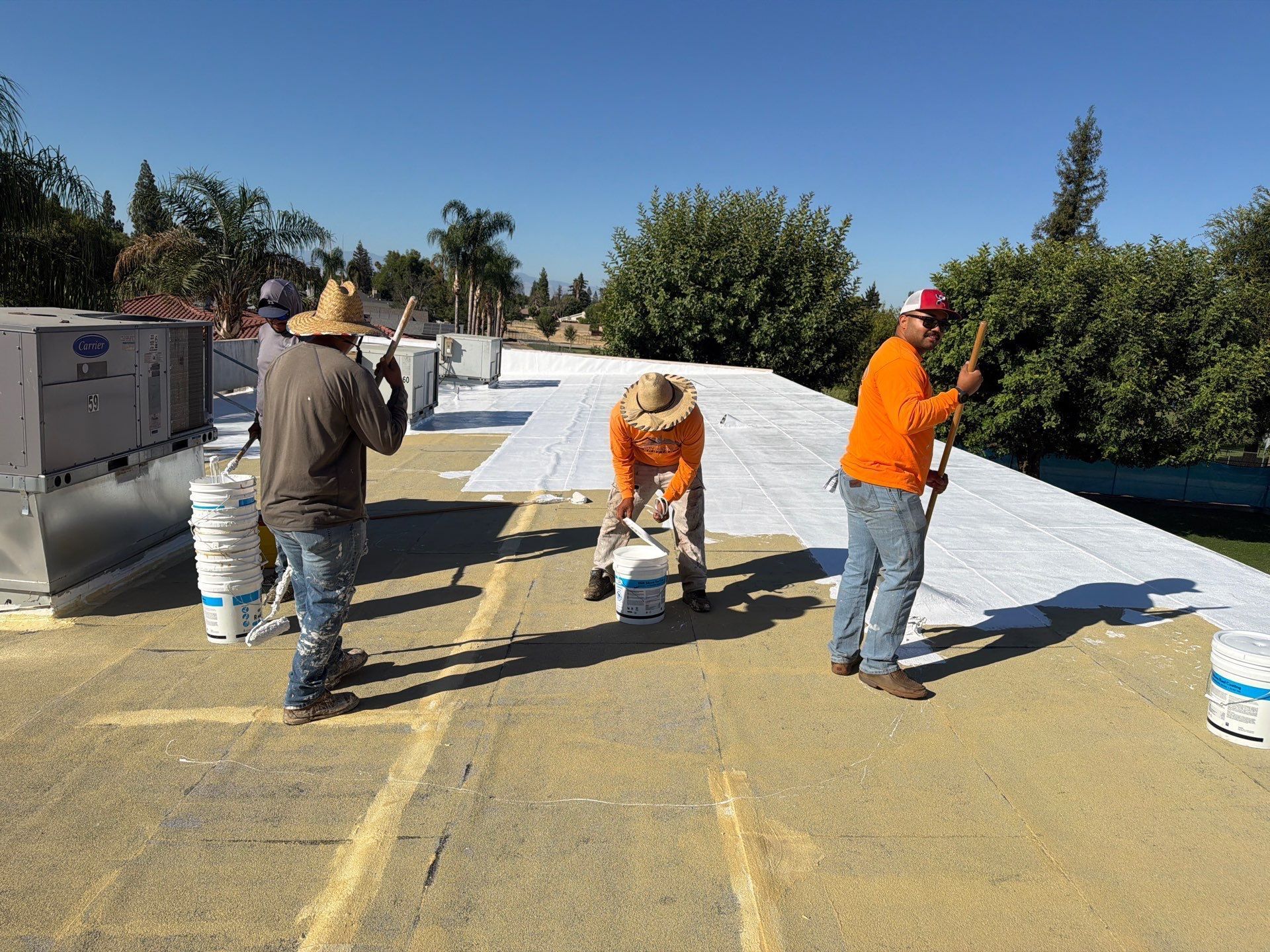 Workers installing roofing on a flat rooftop, with white underlayment and buckets in bright sunlight