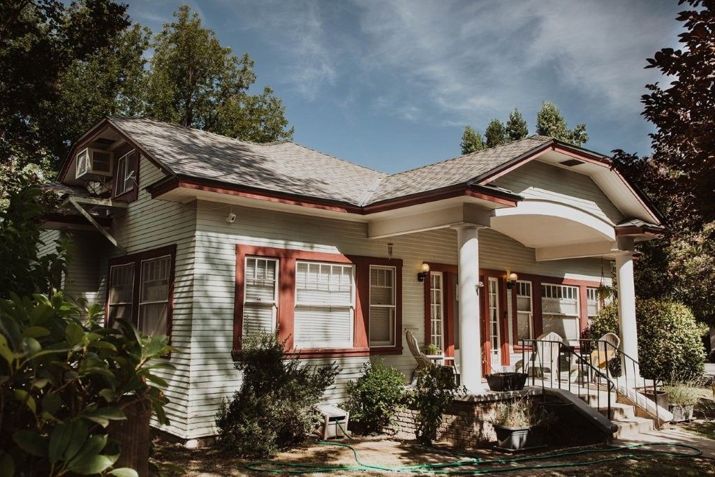 White bungalow with red trim and porch, surrounded by lush greenery under a cloudy sky