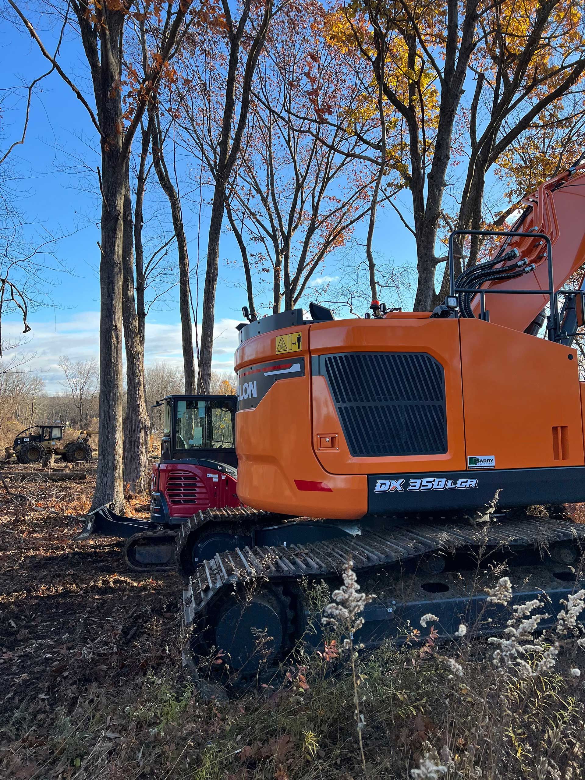 orange excavator and red tractor clearing trees in a wooded area on a sunny day