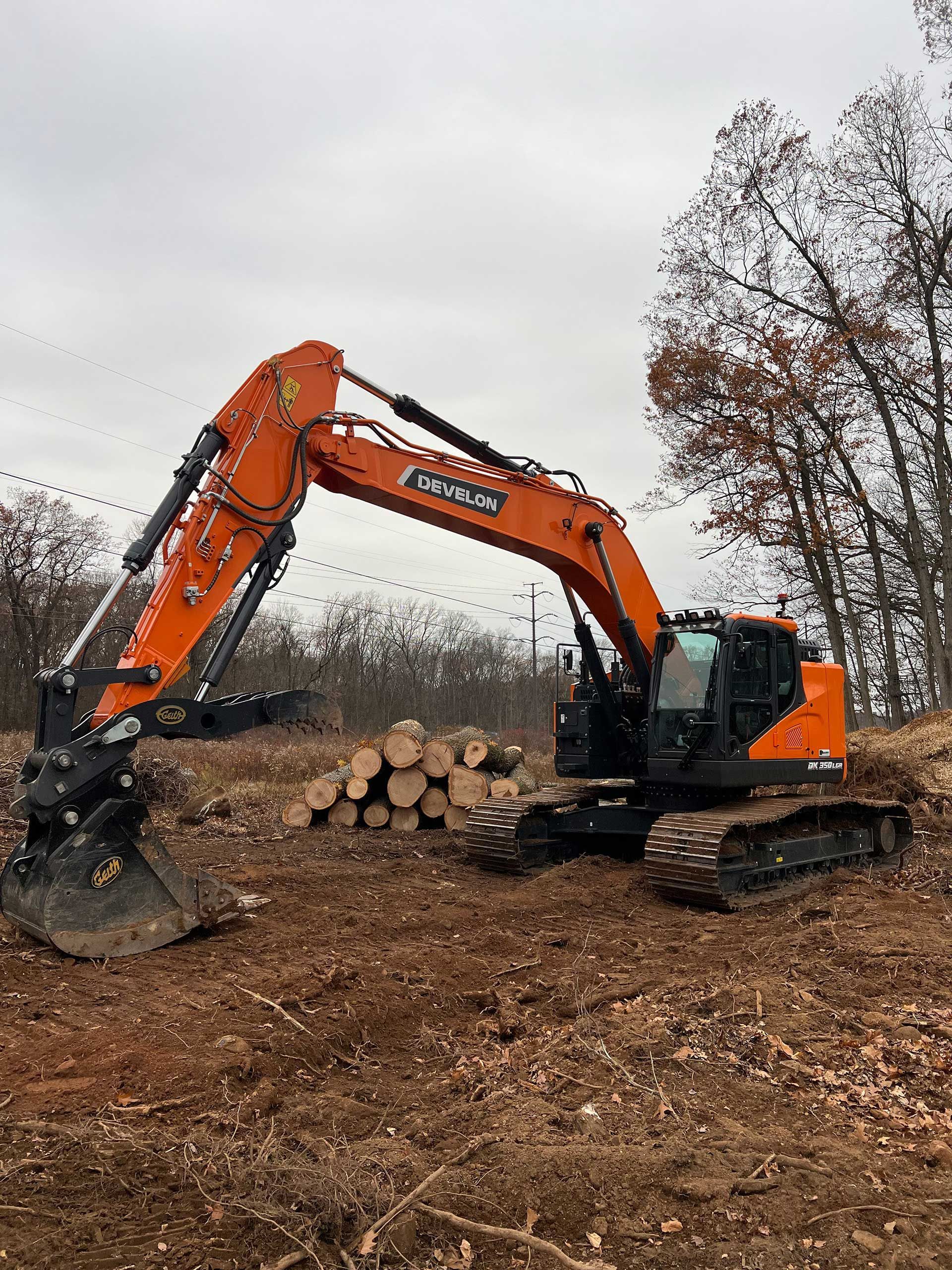 orange excavator on dirt next to a pile of logs, under an overcast sky