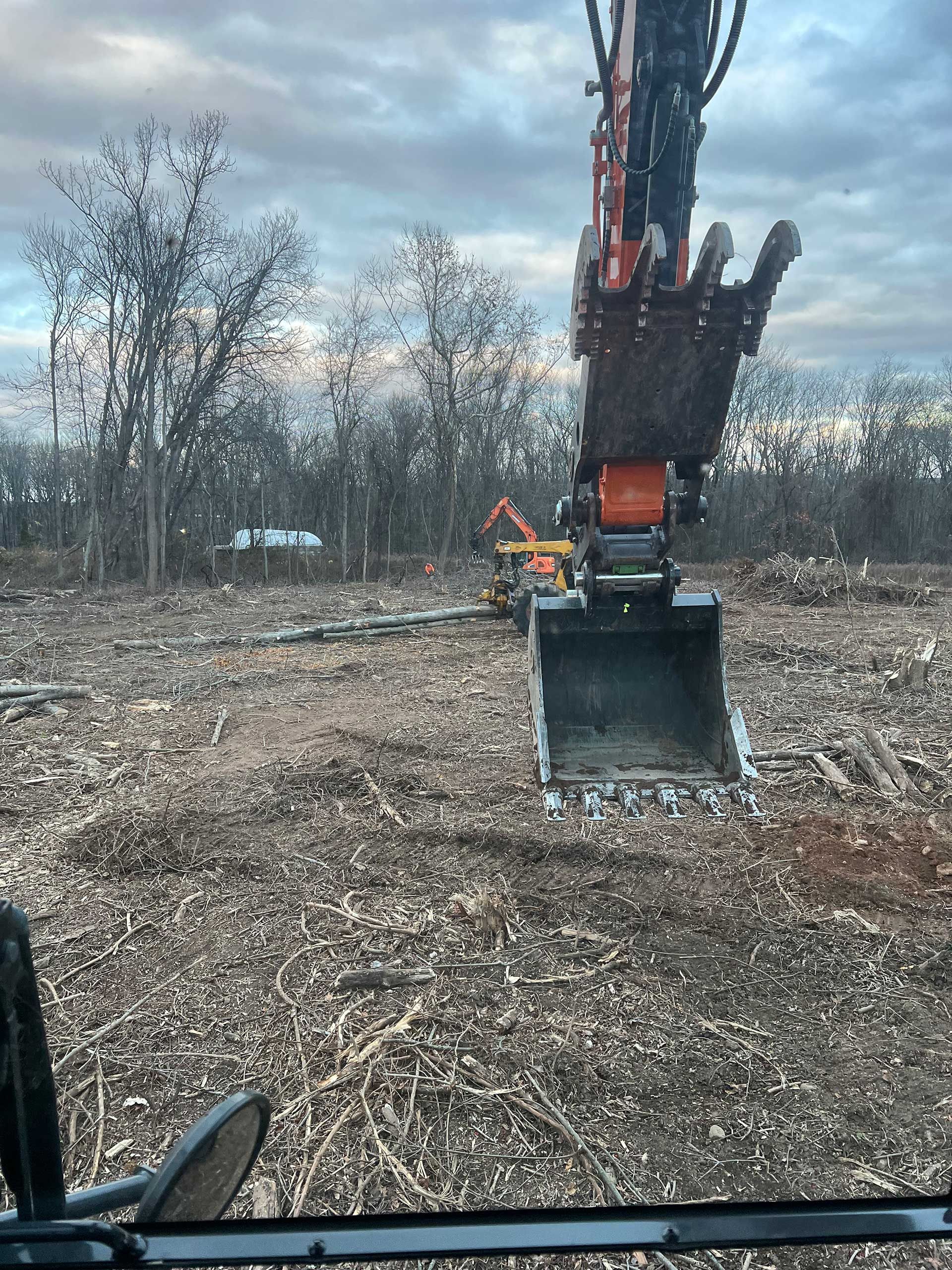 excavator with open bucket in clearing, other machinery in background, under cloudy sky