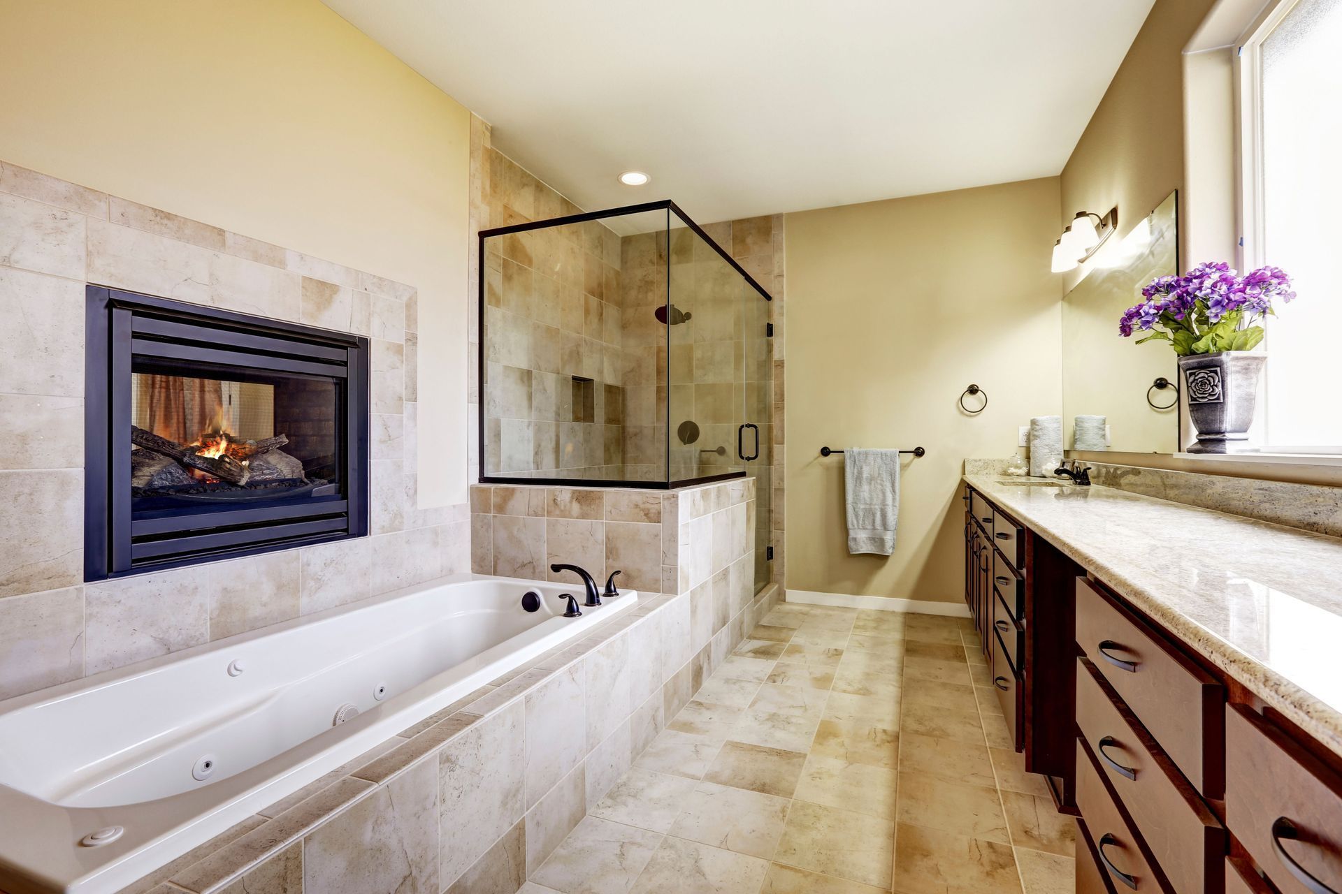 Spacious beige bathroom with a soaking tub, glass shower, fireplace, and vanity with purple flowers.