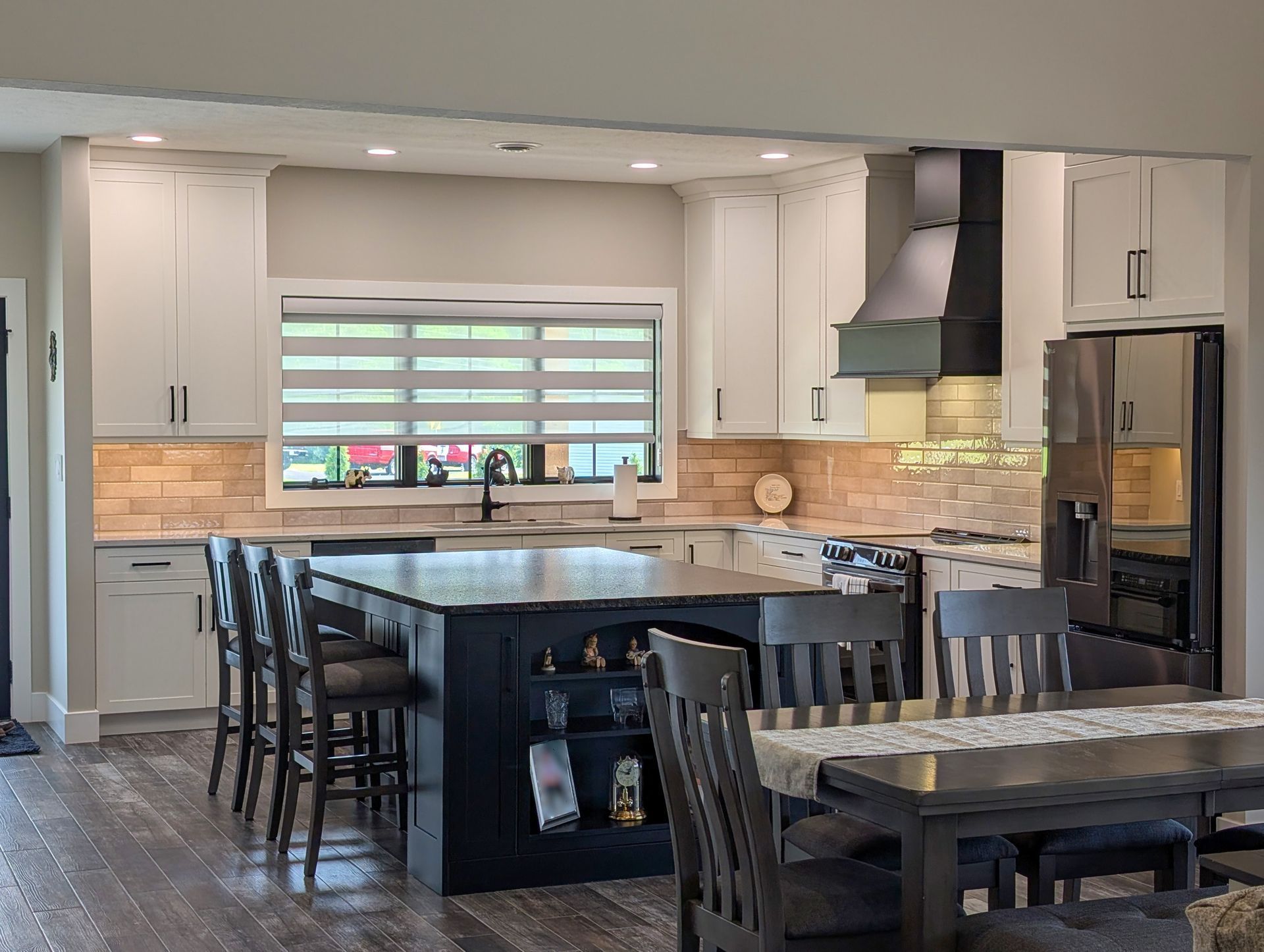 Modern kitchen with white cabinets, dark island, bar stools, dining table, and large window with blinds